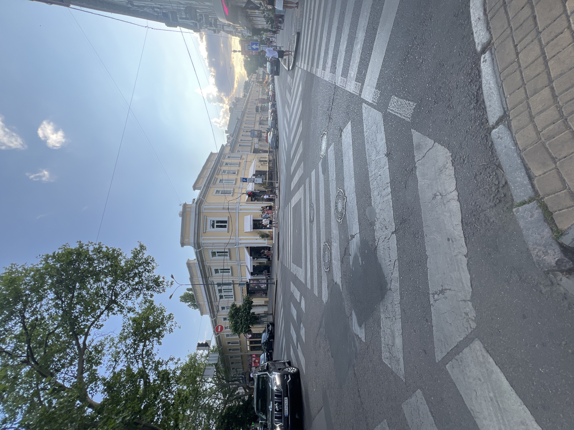 This image depicts a wide street intersection in Odesa, Ukraine, with pedestrian crosswalks prominently featured. The architecture suggests a European city, with buildings of light yellow and cream hues lining the street, some featuring awnings over storefronts. Trees, including a large one with lush green foliage on the left, provide shade. The sky is a clear blue with scattered clouds, hinting at a pleasant day. A dark SUV is stopped at the crosswalk on the left, while several people are seen walking and standing on the sidewalks and at the intersection. The road surface shows wear and tear, with visible cracks and patching on the asphalt. The overall impression is of a bustling urban environment.