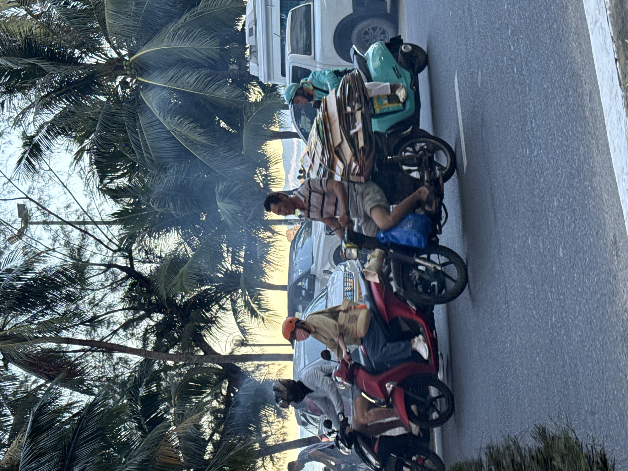 The image captures a street scene in Nha Trang, Vietnam, likely near a coastal area given the presence of palm trees and the soft, hazy light of dusk. In the foreground, several motorbikes are navigating a road. One red motorbike carries a person wearing a tan top, blue pants, an orange helmet, and a face mask. Another motorbike, a teal color, is laden with a large stack of cardboard. The rider on this motorbike wears a teal jacket and helmet, and white shoes. Smoke or exhaust is visible, billowing from behind these vehicles, obscuring parts of the background. Behind the motorbikes, cars and a white truck are visible on the road, and further back, glimpses of palm trees and the sky suggest an outdoor setting. The overall impression is a snapshot of everyday urban life with a tropical backdrop.