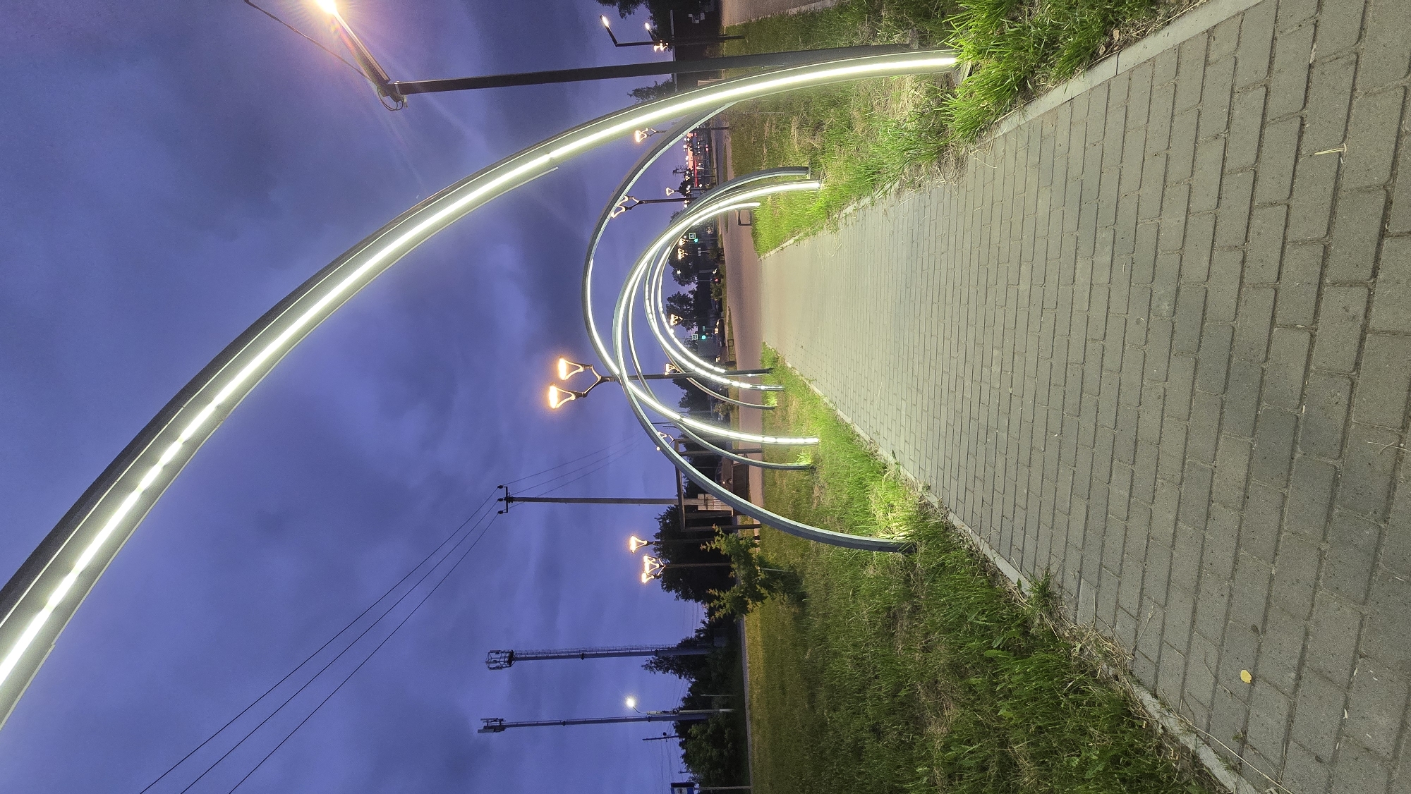 The image depicts a pedestrian walkway at dusk in Chaikovsky, Russia. The walkway is paved with grey brick pavers and is lined with grass on either side. A series of illuminated archways, featuring white LED strip lights, line the walkway, creating a tunnel-like effect. Beyond the archways, the sky is dark blue with some clouds. Traditional streetlights with orange bulbs are visible in the background, as well as utility poles and cell towers. The overall scene is one of urban infrastructure enhanced by modern lighting. There are no people or animals visible in the image.