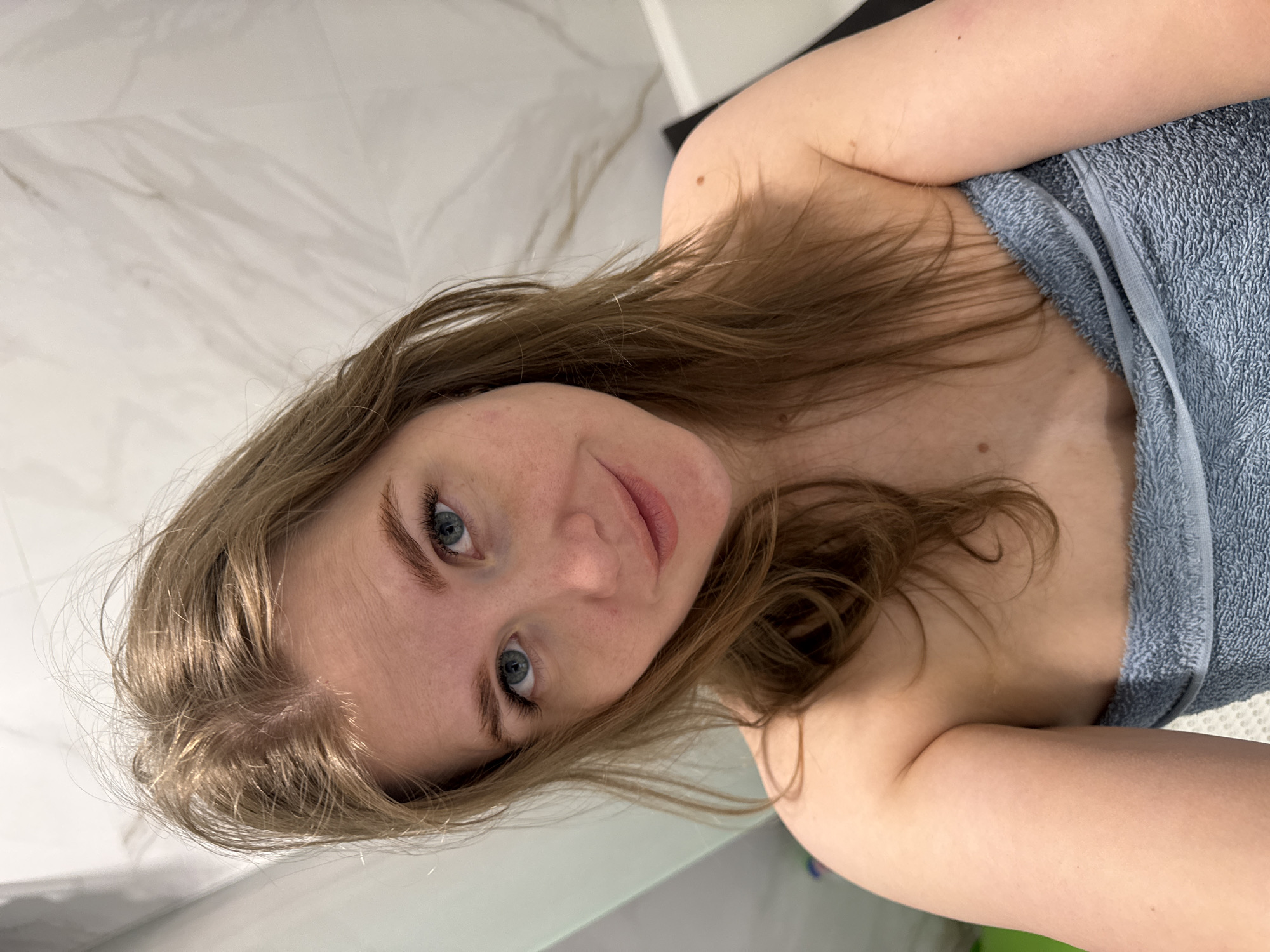 A woman with light brown hair and blue eyes is posing indoors in what appears to be a bathroom. She is wrapped in a blue towel, partially exposing her shoulder and chest. The background consists of white marble-patterned walls. The location appears to be Ферма, Russia.