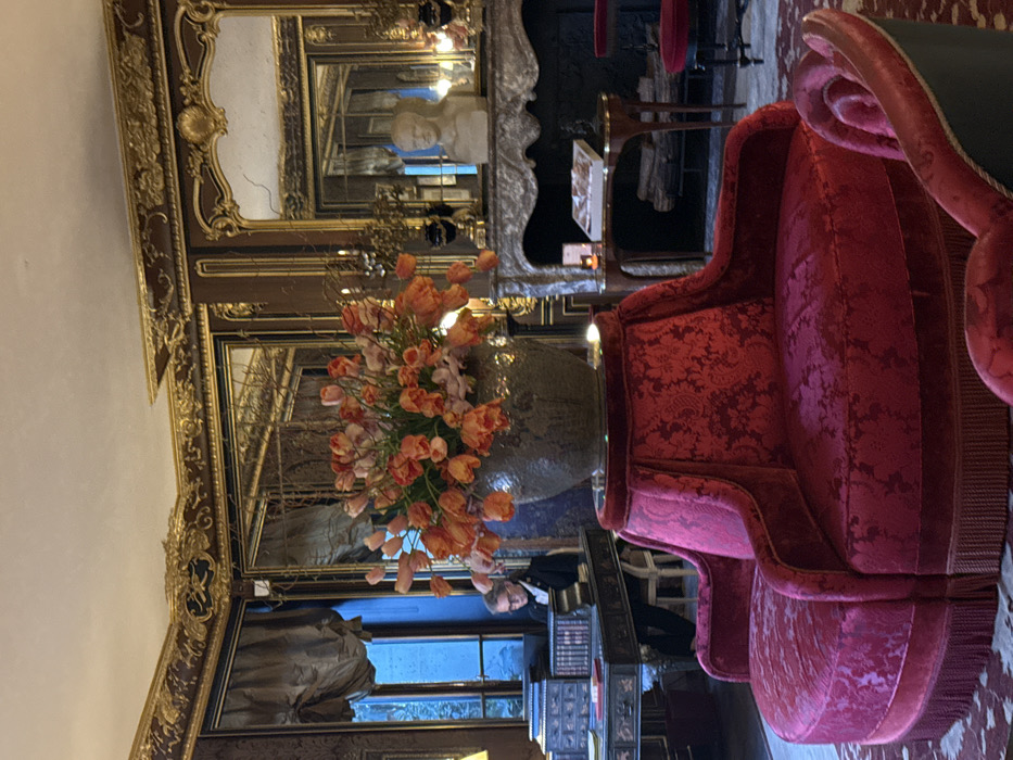 The image depicts an opulent interior scene within a lounge or reception area, identified as **Le Bristol Paris, France**. The room features dark wood paneling with extensive gold gilding on cornices and wall trim, creating an elaborate frame for large mirrors that reflect parts of the room. The ceiling is white with matching gilded molding.
In the foreground, a large circular seating unit covered in deep red velvet with a prominent damask pattern and a fringed base occupies the center-right. It has multiple sections, forming a conversation pit style. On a circular velvet-covered stand positioned directly in front of this seating, a large, dark gray vase holds a vibrant arrangement of numerous orange and some pink tulips.
To the left, against a window, a dark wooden writing desk with multiple drawers and stacked books is visible. A person, appearing to be a staff member or concierge, is seated at this desk, wearing dark attire and glasses, focused on an item on the desk. Dark drapes frame the window, which shows a muted blue light from outside.
To the right, a marble fireplace with an intricately carved, gilded mantelpiece is a central feature. On the mantel, a white male bust statue is flanked by two dark urns with gilded elements. A small, elegant wooden side table with a single lit candle and a book is placed next to the fireplace. A smaller red stool or chair is partially visible further right. The floor is covered with a patterned red rug. The overall lighting is dim and ambient, highlighting the room's luxurious and historic decor.