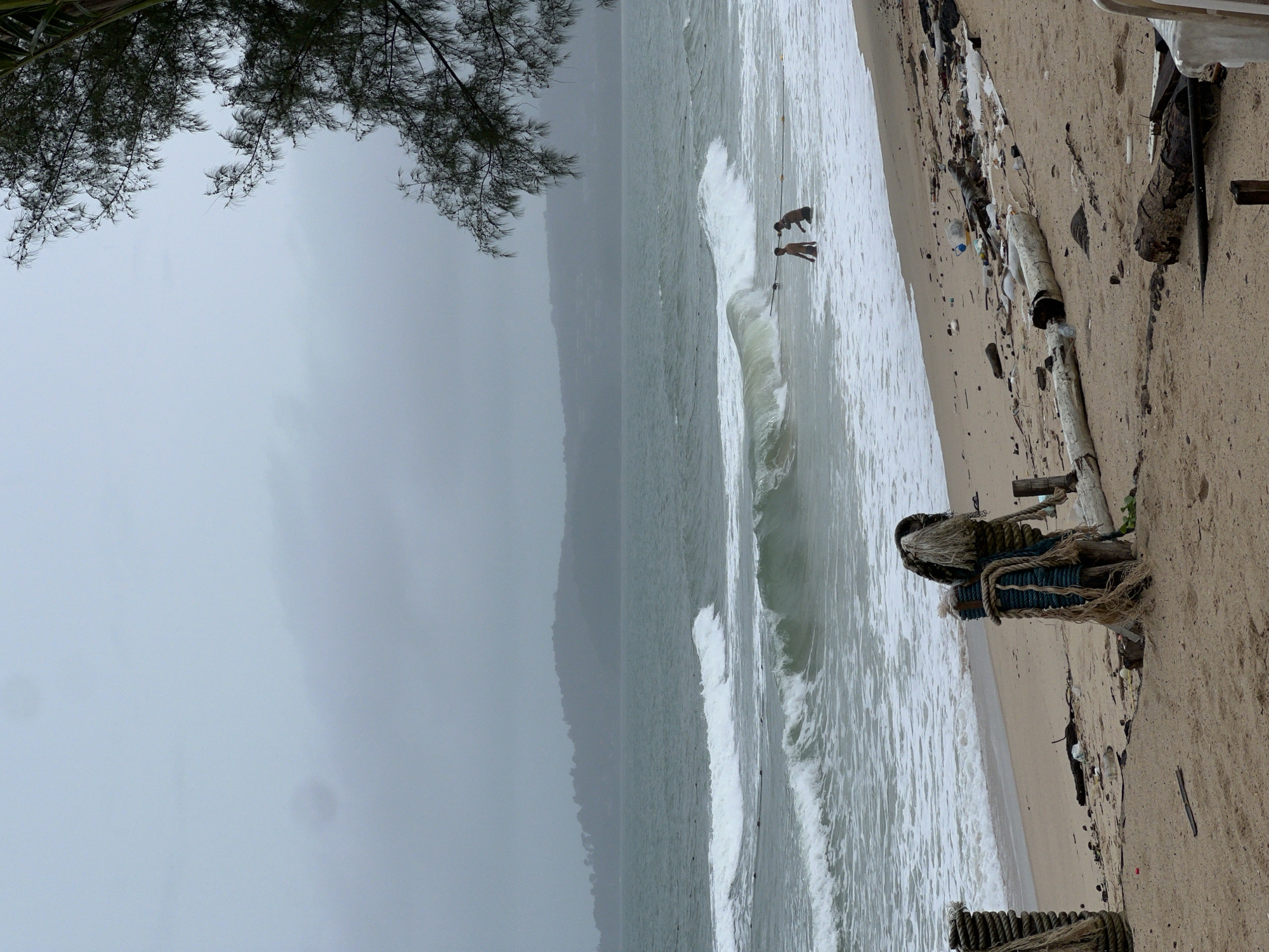 The image depicts a beach scene on an overcast day, captured with a vertical orientation (sky at the top, ocean and beach in the lower half when rotated correctly). In the upper left corner, dark, pine-like tree branches frame a portion of the dull gray sky.
The ocean, rendered in shades of gray and green, exhibits active waves, with a prominent wave breaking in the middle ground. Two individuals, appearing to be children or young adults, are visible in the water, close to the shore, interacting with the breaking waves. One figure is partially submerged or leaning into a wave, while the other stands nearby.
The sandy beach occupies the lower right portion of the frame and is littered with various forms of debris. Prominent among these are several large pieces of driftwood or logs. In the immediate foreground, remnants of what appears to be fishing gear or nautical ropes, tangled and worn, are secured to wooden posts or debris. Other smaller pieces of litter, including plastic bottles, are scattered across the sand.
A hazy, dark landmass, possibly a distant island or coastline, is faintly visible across the water in the background, blending into the muted sky. The overall weather appears cloudy and possibly breezy, contributing to the choppy water and the subdued color palette dominated by grays and greens. No visible text is present in the image.
