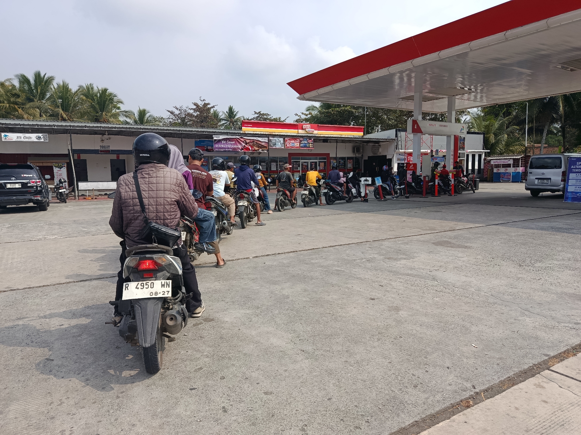 A line of motorcycles and their riders are waiting at a Pertamina gas station in Desa Rawajaya, Indonesia. The scene is outdoors on a paved surface. The gas station has a red and white canopy with a white ceiling. To the left, there is a building with a tiled roof and some trees visible behind it. Several motorcycles are lined up, with the closest one in the foreground facing away from the camera. People are seated on their motorcycles, some wearing helmets. The sky is partly cloudy. Signs are visible on the buildings, including one that reads "TOILET". Another sign in the distance shows a large advertisement with red text and images of food items. A silver van is parked to the right. The ground is concrete with expansion joints.