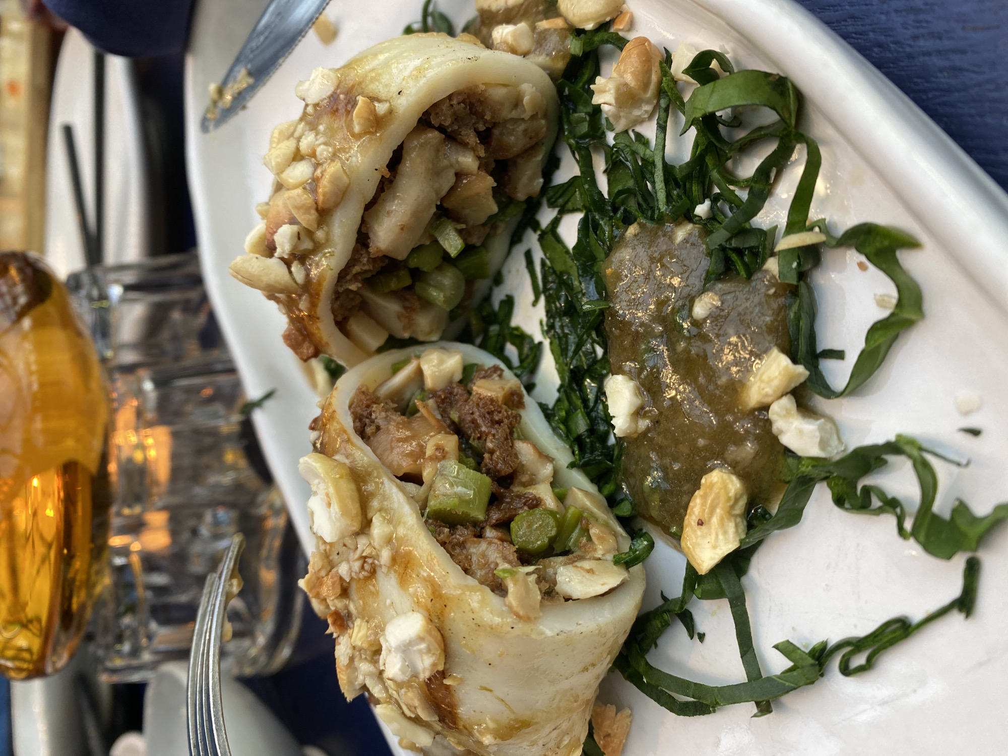 A close-up shows a white rectangular plate with three stuffed squid. The squid are filled with a mixture of meat, vegetables and are topped with chopped nuts. Beside the squid is a dollop of dark sauce surrounded by chopped greens, also topped with chopped nuts. A fork and knife are on the left side of the frame. A glass with a yellow liquid is on the left side of the plate, partially obscuring the plate. The plate appears to be set on a blue surface. The location is not determinable from the image.