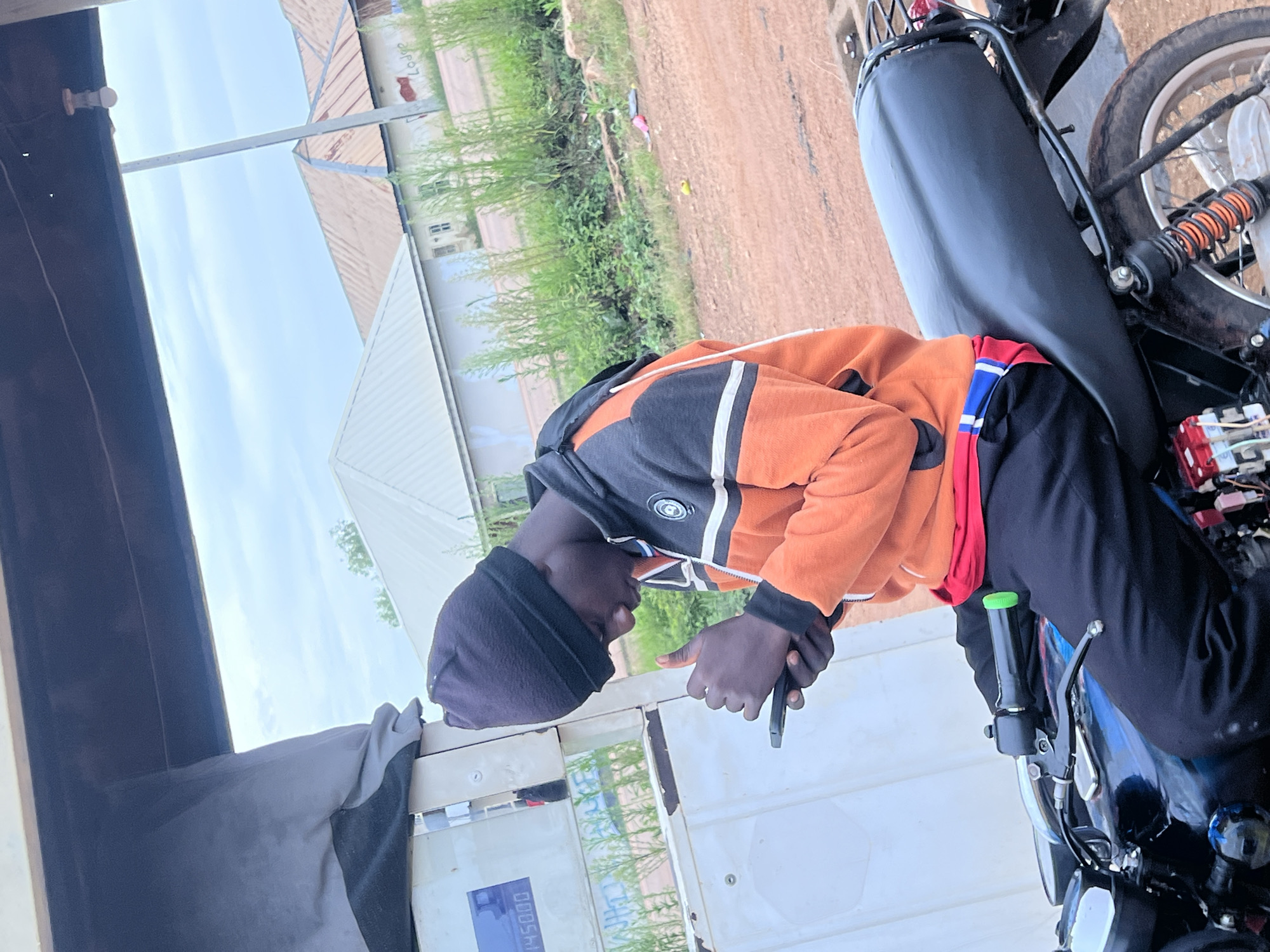 A young Black male, wearing a black beanie, an orange and black hooded jacket with white stripes, and black pants, is sitting on a black motorcycle. He is holding a smartphone in his left hand and appears to be interacting with it. The motorcycle is parked next to a fuel pump with a digital display showing "1450.00". In the background, there are several buildings with corrugated metal roofs and a cloudy sky. The ground is a dusty, reddish-brown dirt road. The scene is located in Durimi, Nigeria.