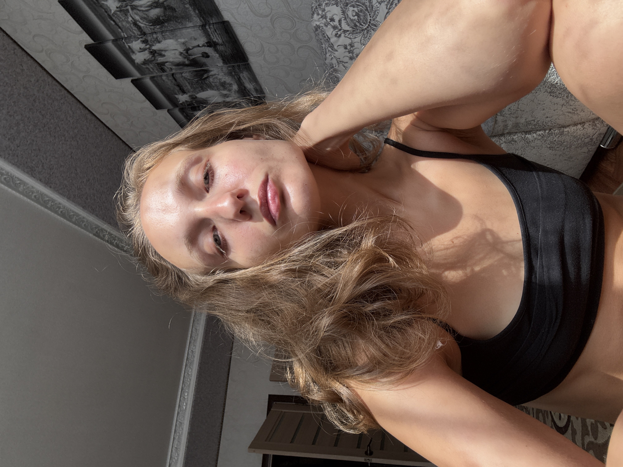 A young woman with blonde, wavy hair is captured in a close-up, frontal shot. She is wearing a black sports bra and has her left arm resting on her neck. Sunlight is casting strong shadows across her face and body. In the background, a door is visible to the left, and a triptych artwork depicting horses running is on the wall to the right. The setting appears to be an indoor space, possibly a bedroom, with textured wallpaper and a patterned surface on the right side. The image does not provide any specific context to suggest it is located in Kuyeda, Russia.