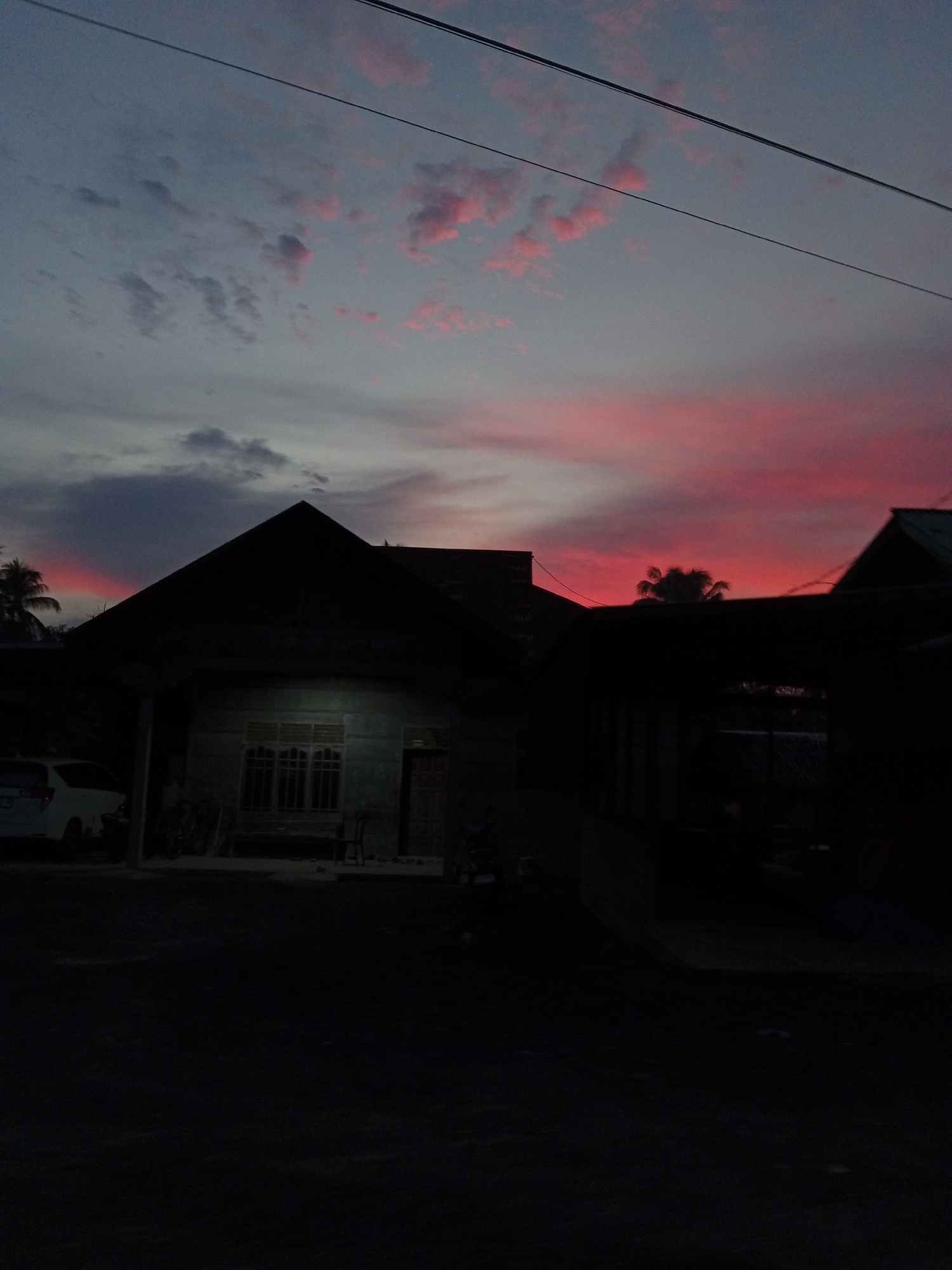 The image depicts a twilight scene with a vibrant pink and grey sky, possibly indicating sunset or sunrise. Silhouetted against the sky are the dark shapes of rooftops and a palm tree. In the foreground, the dark outlines of houses and a vehicle are visible, with some faint illumination suggesting ambient light or a street light. The scene appears to be an outdoor residential area. The overall impression is of a peaceful, atmospheric moment in a town. It is understood that this image was taken in Blang Mee, Indonesia.