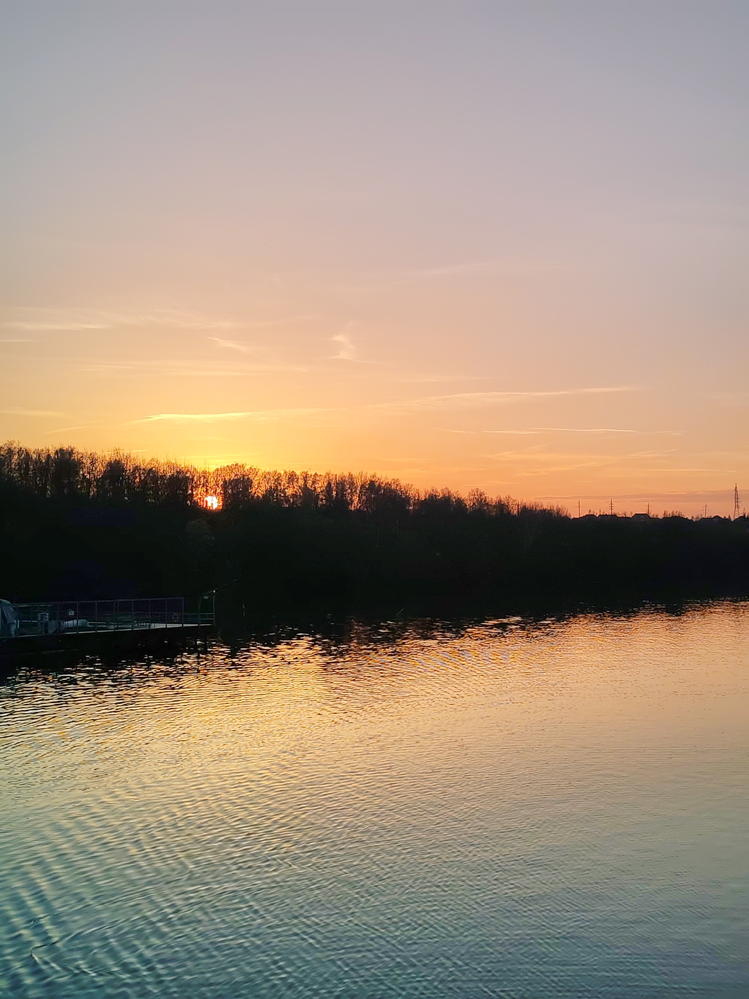 The image shows a sunset over a body of water, possibly a river or lake. A line of trees runs along the horizon, silhouetted against the setting sun. To the left, there appears to be a dock or pier extending into the water. The sky is a gradient of colors, ranging from orange near the horizon to lighter shades of pink and blue above. The water reflects the colors of the sky, creating a shimmering effect. The location is likely in the city of Сигнал Снт, Russia, based on the metadata provided. There are no people visible in the image.