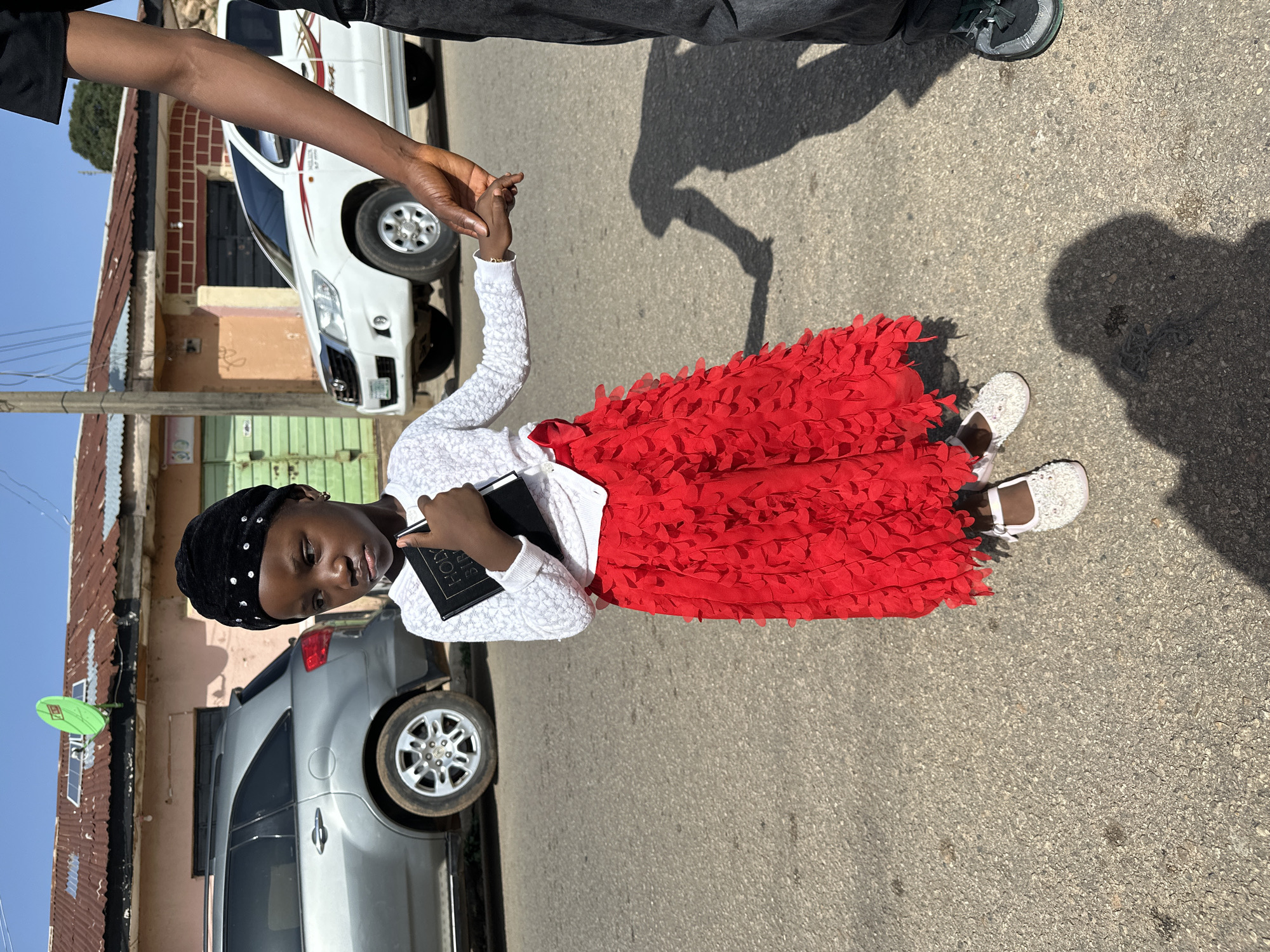 A child wearing a white sweater, red skirt, black head covering, and white shoes is holding a black book and holding hands with an adult in Unknown, Unknown. They are standing on a paved street in front of buildings and parked cars. The sky is blue.