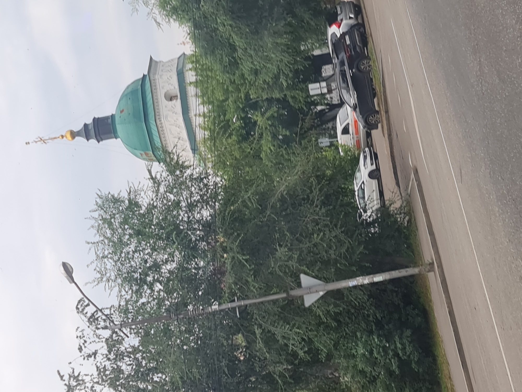 The image shows a street scene in Irkutsk, Russia, with a prominent church dome visible in the background. A tall street lamp with a triangular sign is in the foreground, and several cars are parked or driving on the road. Trees line the street, partially obscuring the view of the church and some of the cars. The sky is overcast. The church appears to be a white building with a green dome topped with a gold cross.