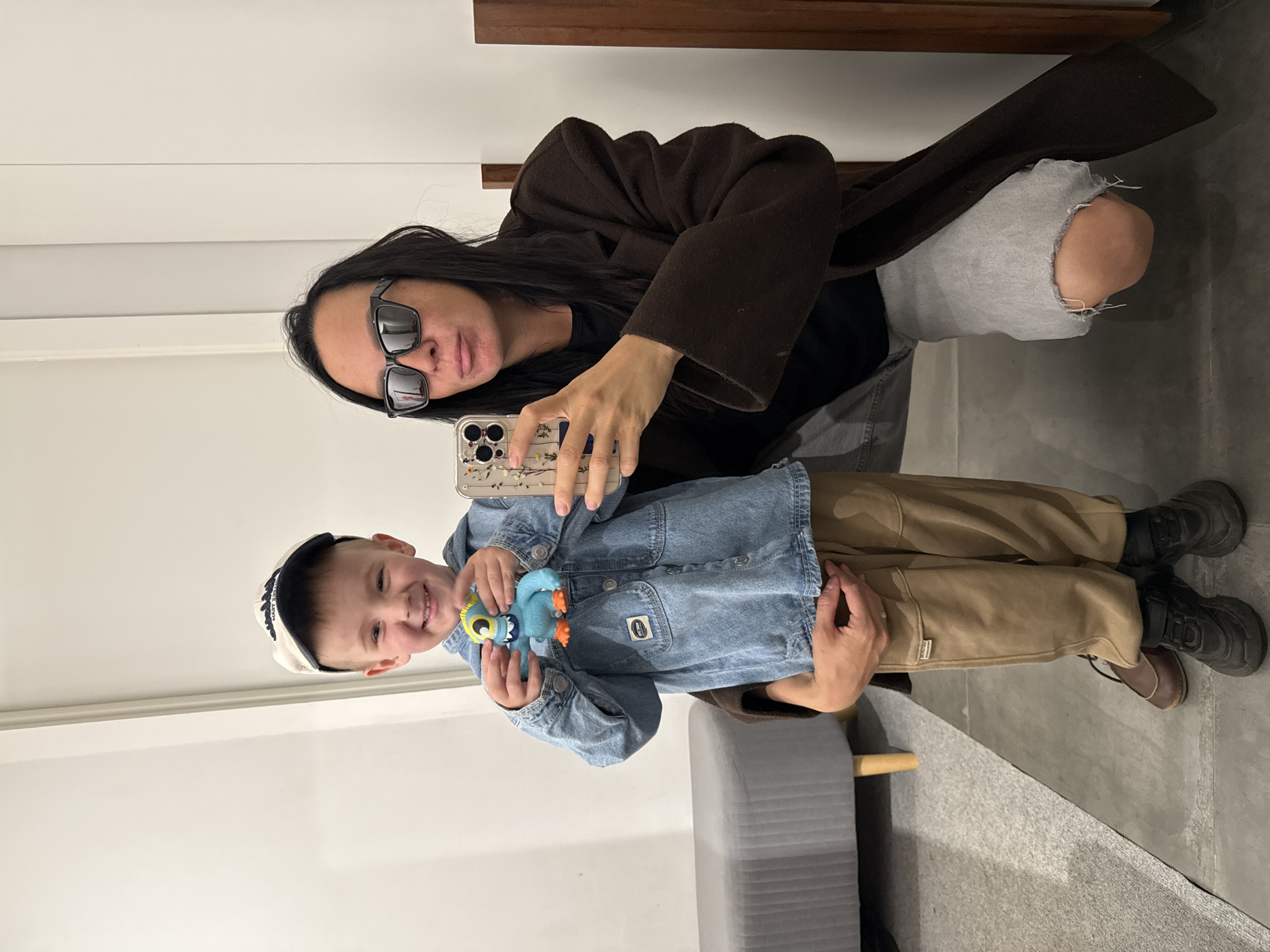 In an indoor environment, a young boy wearing a denim jacket and a baseball cap, holding a blue toy, stands in front of an adult. The adult, dressed in a dark coat and sunglasses, is taking a selfie with the boy, holding up a smartphone. The background consists of a white wall and a grey upholstered bench. The floor is tiled. The image appears to be taken in a retail setting or a home. The specific location within the city of Chaikovsky, Russia, cannot be determined from the visual information provided.