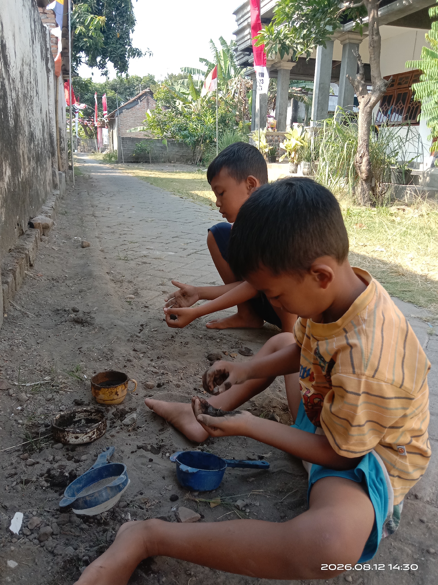 The image depicts two young boys playing in the dirt on a paved walkway in Banyuarang, Indonesia. The boys, of Asian descent and appearing to be under the age of ten, are engaged in activity with the soil. The boy in the foreground wears a yellow striped t-shirt and blue shorts, and is focused on the dirt in his hands. The boy behind him, wearing a dark shirt, also appears to be interacting with the ground. Scattered around them are several small objects, including two metal cups, one yellow and one white, a blue plastic sieve, and a blue plastic bowl. The environment is outdoors, with a stone wall to the left, trees and vegetation, and a building in the background. Indonesian flags are visible on poles in the background, suggesting a local celebration or event. The lighting indicates it is daytime, with shadows present. A date and time stamp, "2026.08.12 14:30", is overlaid on the lower right corner of the image.