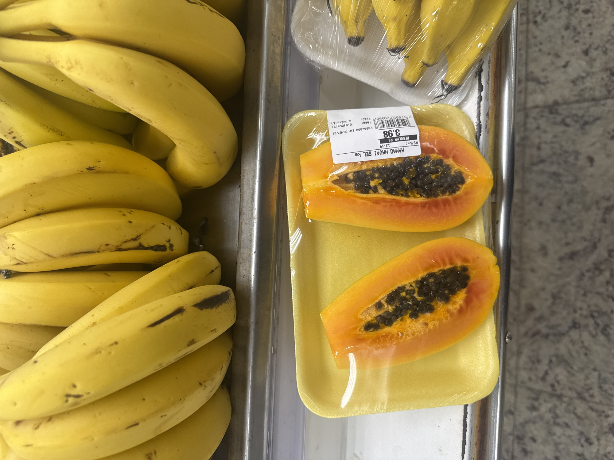 Two halves of a papaya are displayed on a yellow Styrofoam tray, wrapped in clear plastic. The papaya flesh is orange with numerous small, black seeds in the center. The halves are positioned side-by-side, with the cut surfaces facing upwards. A white sticker with text and a barcode is attached to the plastic wrap. In the background, a pile of ripe yellow bananas is visible, also wrapped in plastic. The fruits are on a shelf in what appears to be a grocery store or market. The lighting suggests an indoor environment, likely a refrigerated display case, given the condensation on the plastic wrapping of the bananas. The floor or surface beneath the shelf is visible, with a speckled grey pattern. The scene does not contain any people.