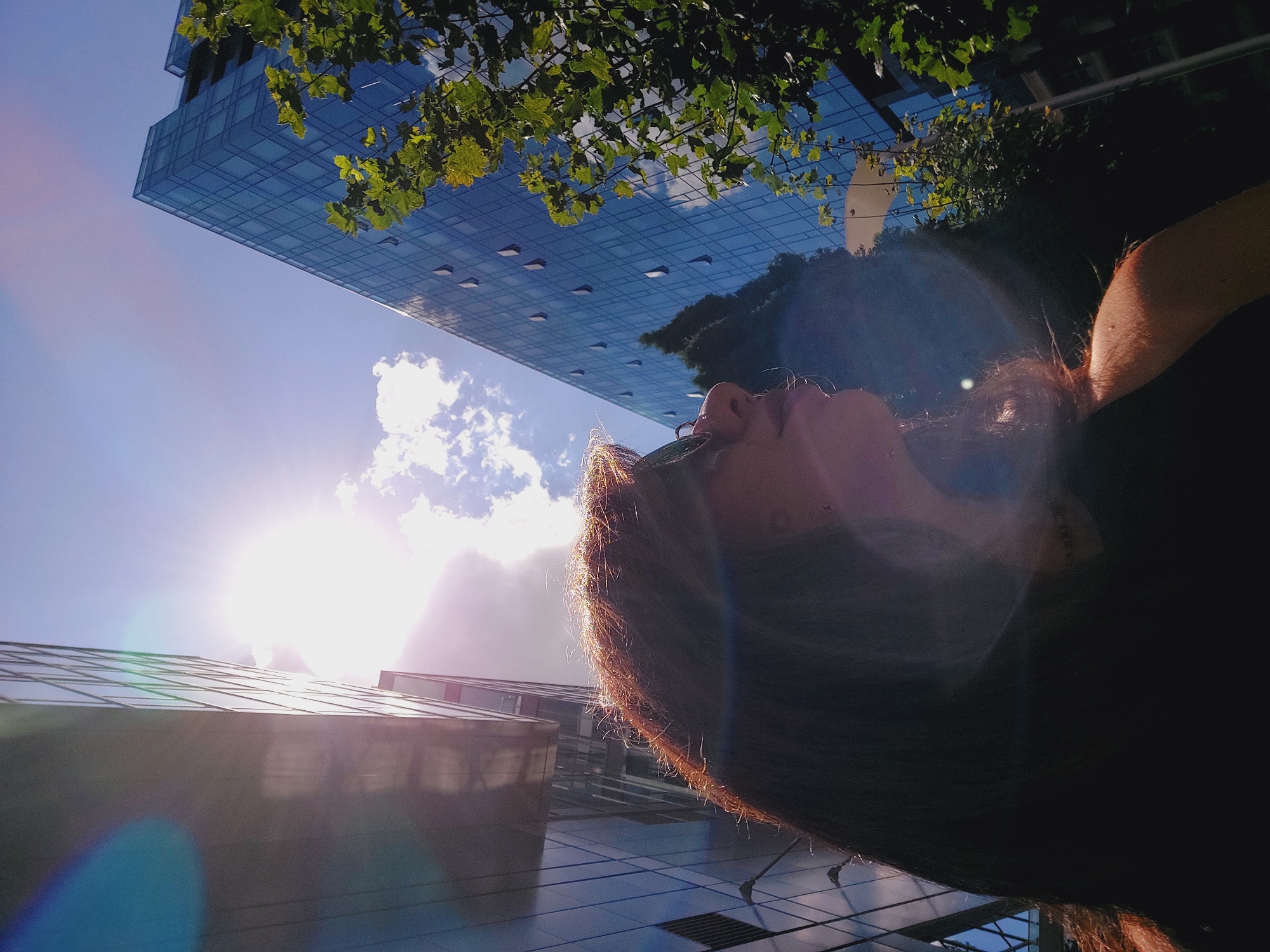 A low-angle, close-up shot captures a woman in profile, looking upwards towards the sun. Her long, auburn hair is highlighted by the bright sunlight, and she wears dark sunglasses. The background consists of modern glass buildings and green trees, suggesting an urban environment. The sun is intensely bright, creating lens flares and overexposed areas, obscuring details in the sky and on the buildings. The left building appears darker and has a reflective surface, while the building on the right is primarily blue glass. The image has a hazy, sun-drenched quality.