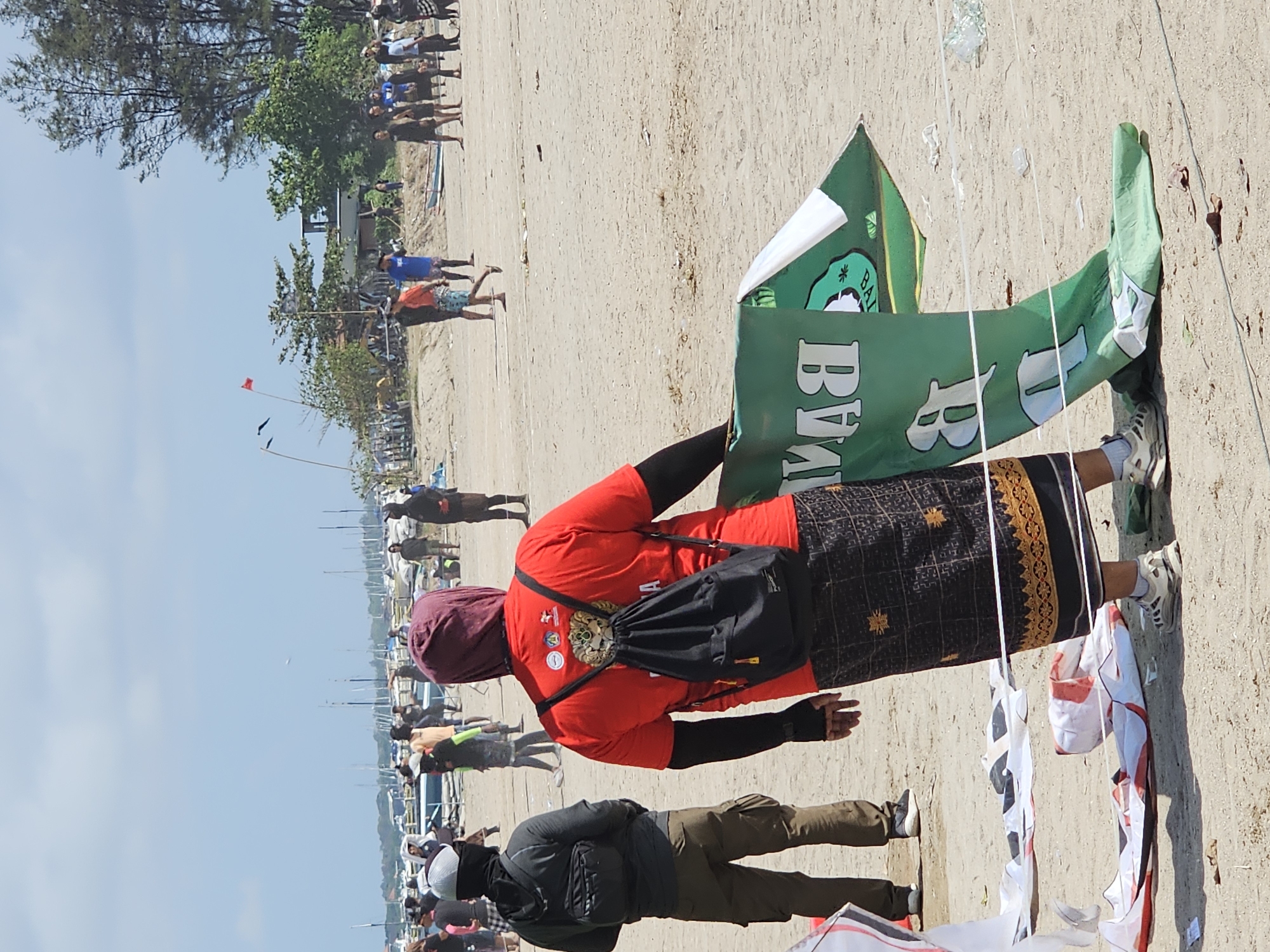 The image depicts a beach scene in Denpasar, Indonesia, likely during a kite festival given the presence of numerous kites in various states of assembly and flight, as well as a large crowd of people. In the foreground, a person wearing a red shirt, a purple headscarf, and a black backpack stands holding a large green banner. The banner, partially unrolled, displays white lettering. To the left, another individual is visible, wearing a grey hoodie and cargo pants, also facing away from the camera. The beach is sandy, with the ocean and numerous boats visible in the background. The sky is clear and blue, with a few wispy clouds. Several other people are scattered across the beach, some appearing to be participating in or observing the kite activities. The overall atmosphere suggests a lively outdoor event.