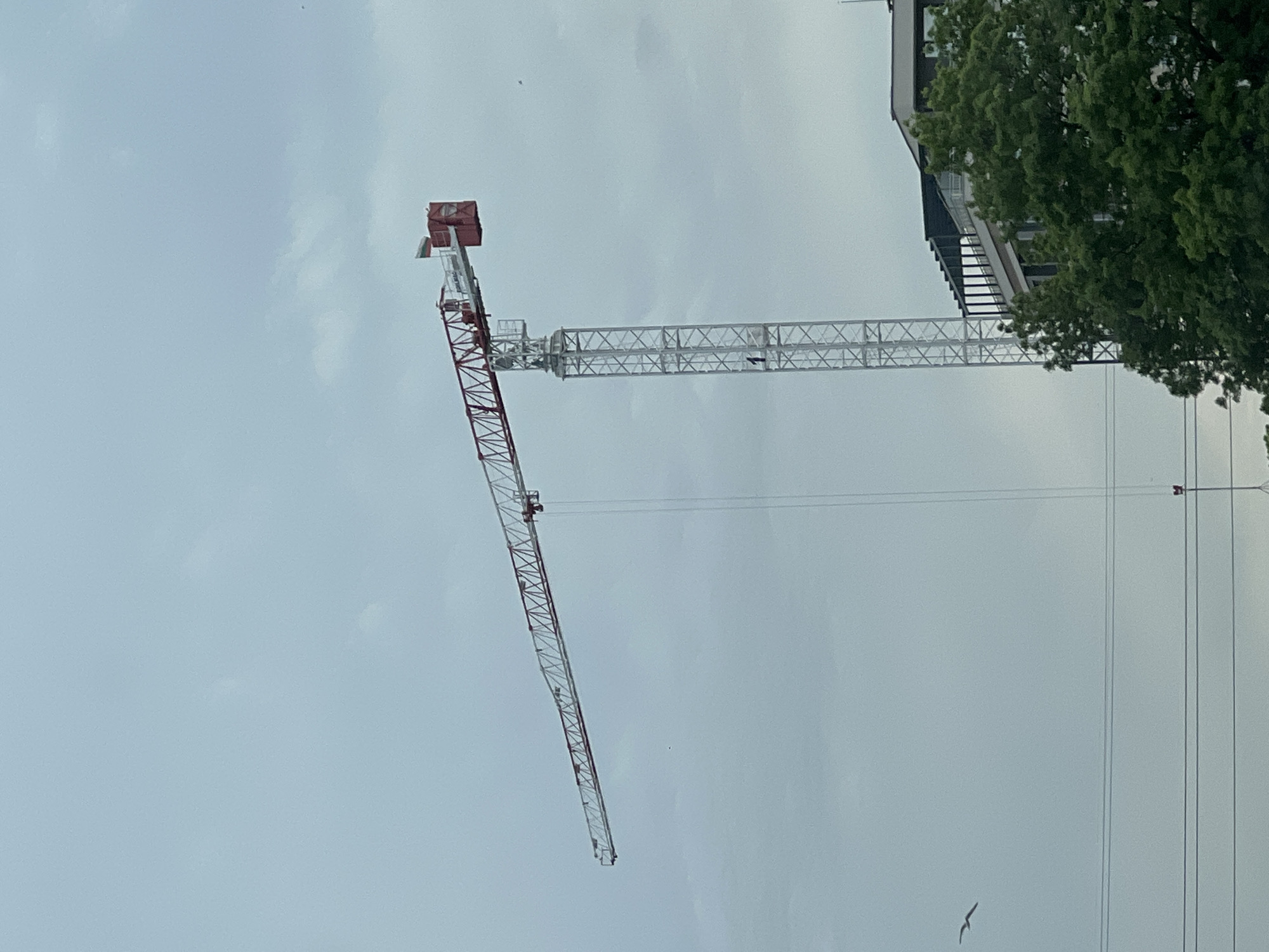 The image depicts a construction crane against a cloudy sky. The crane is red and white, with a horizontal boom extending to the right and a vertical arm extending downward. A building is partially visible behind the crane, with a tree partially obscuring it. Power lines are visible to the right of the frame. A bird flies near the bottom of the image. The location is Burgas, Bulgaria.