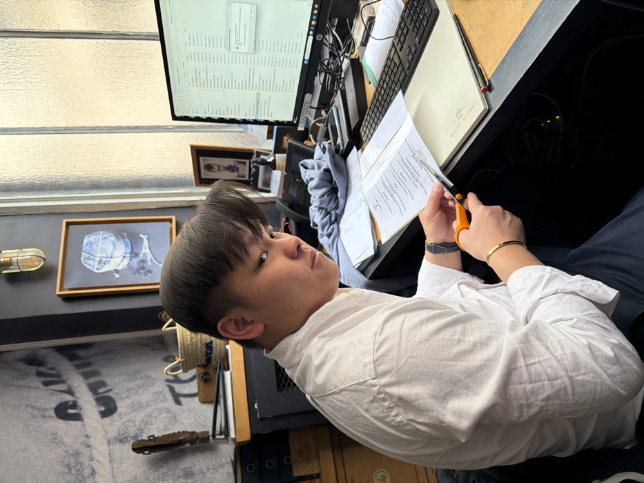 An East Asian male, estimated to be in his late 20s to early 30s, is seated at a dark-colored desk in what appears to be an office or workspace. He has short, dark hair with a distinct fringe and is wearing a white long-sleeved collared shirt and dark trousers. On his left wrist, he wears a dark watch, and on his right, a thin gold-colored bracelet. He is actively cutting a white piece of paper, possibly a menu with the word "MENU" visible, using orange-handled scissors. His gaze is directed towards the viewer with a neutral expression.
The desk area to his right features a black desktop computer monitor displaying a file explorer window with numerous folders and a small alert dialog box in the upper right corner, indicating a potential issue with a local drive ("H:\..."). To the left of the monitor are a black desk phone and a crumpled light blue sweatshirt. A light-colored, possibly leather-bound, folder or notebook with "MENU" imprinted on its cover lies adjacent to the paper being cut. A black keyboard and mouse are also visible on the desk. Below the desk, cables and small green indicator lights are visible.
Behind the subject, a dark grey wall is adorned with a large, faded mural of light-colored abstract shapes and text. A framed artwork hangs on this wall, depicting a light blue spherical object (resembling a hot air balloon) with a representation of the Eiffel Tower beneath it. This artwork strongly suggests the location is in Paris, France. Above the artwork, an industrial-style wall light fixture with a metal cage and glass bulb is mounted. A window with textured or frosted glass is visible to the right of the artwork, allowing diffused natural light into the room. On a shelf or cabinet to the subject's left, stacked dark boxes, a woven straw basket, and a decorative wooden object are present.