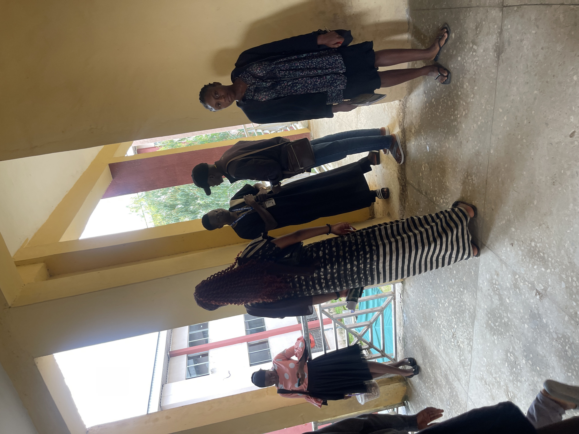 In an outdoor corridor with speckled concrete flooring, several individuals are gathered. The architecture suggests a modern, possibly institutional building, characterized by yellow-painted walls and pillars, and open-air walkways.
On the left, a woman in a pink polka-dot blouse with flared sleeves and a black skirt stands near a railing, looking at her phone. Beside her, a person with long, reddish-brown braided hair, wearing a black and white striped dress, is partially visible from the back.
In the center, two men converse. One, wearing a black robe and a black baseball cap, appears to be speaking, while the other, in a brown jacket and jeans, listens. A brown messenger bag is slung over his shoulder.
To the right, a woman in a dark floral-patterned top and black skirt stands facing forward, holding a document. She wears a dark jacket. Her footwear consists of black flip-flops.
The background reveals a building with red and white accents and windows. Beyond that, lush green foliage suggests a natural environment. The overall scene depicts a casual gathering or encounter in a public space. The location of Jos, Nigeria is not discernible from the image itself.