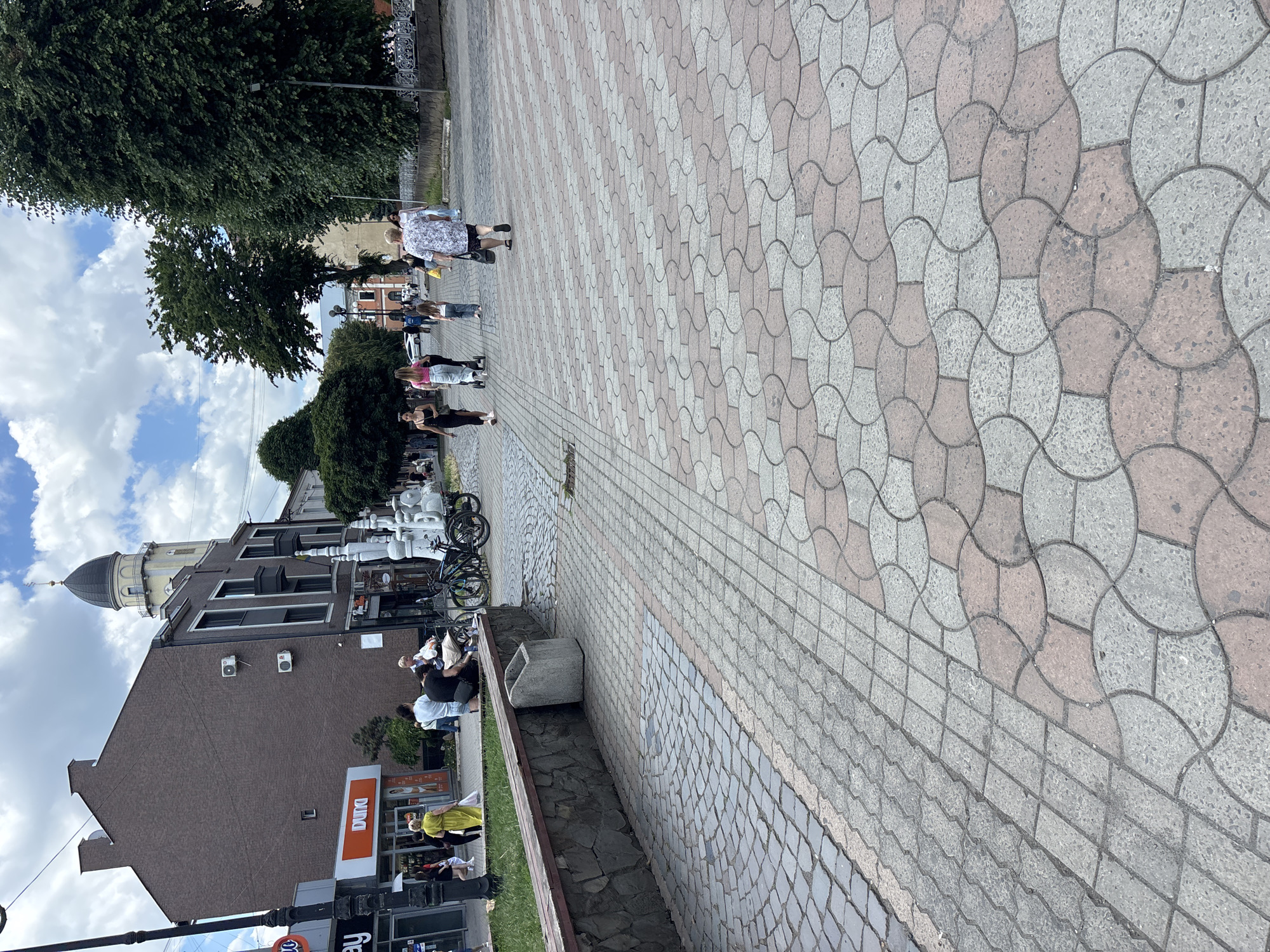 The image depicts a paved pedestrian area in a city. In the foreground, the ground is covered with interlocking stone pavers in a pattern of grey and reddish-brown circular shapes. To the left, a section of grass is bordered by a stone wall. Beyond the wall are buildings, including a brick building with a tall tower topped with a dome and a cross, and a storefront with an orange sign that reads "DUNA". Several people are walking on the paved area, including a woman in a yellow dress, a woman pushing a stroller, and other individuals walking in various directions. Two bicycles are parked near the storefront. In the background, there are trees and a cloudy sky. The scene appears to be a public space within an urban environment, likely a city square or a pedestrian walkway.