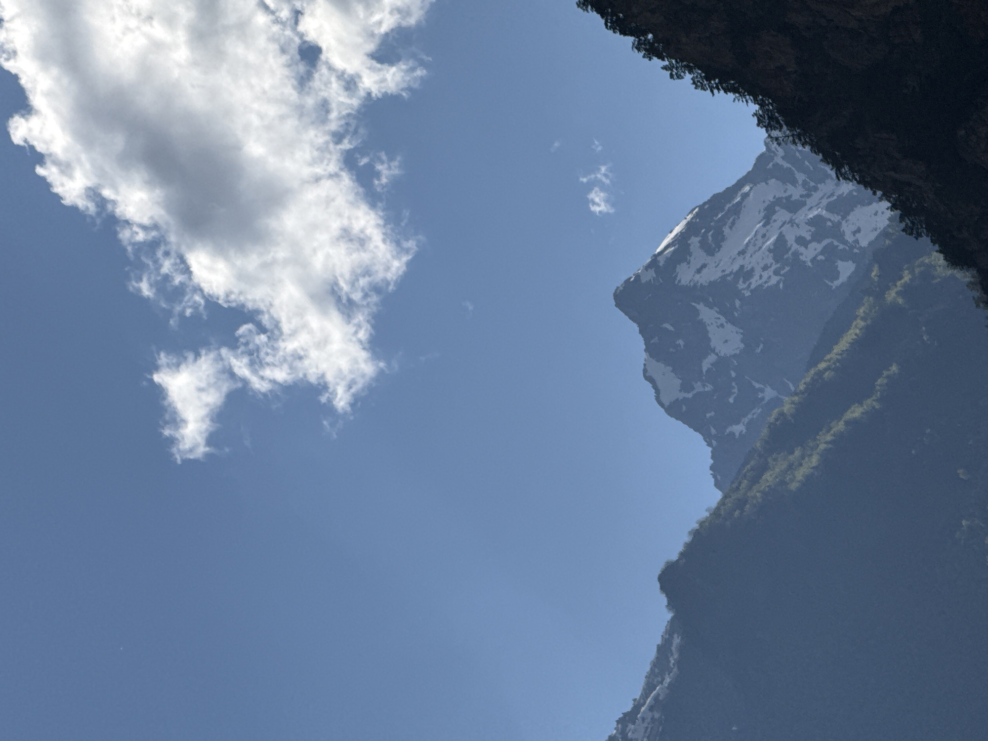 The image depicts a majestic mountain landscape under a clear blue sky with scattered white clouds. In the foreground, the dark, rocky slopes of mountains are visible, densely covered with coniferous trees. The middle ground is dominated by a towering, snow-capped mountain peak, its rugged slopes partially obscured by shadows. To the left, another mountain range, softened by atmospheric haze, is visible, with hints of green vegetation and snow patches. The overall scene conveys a sense of natural grandeur and untouched wilderness, likely located in a mountainous region such as Teberda, Russia, known for its dramatic scenery and alpine environment. There are no people or specific objects of human construction visible in the image.