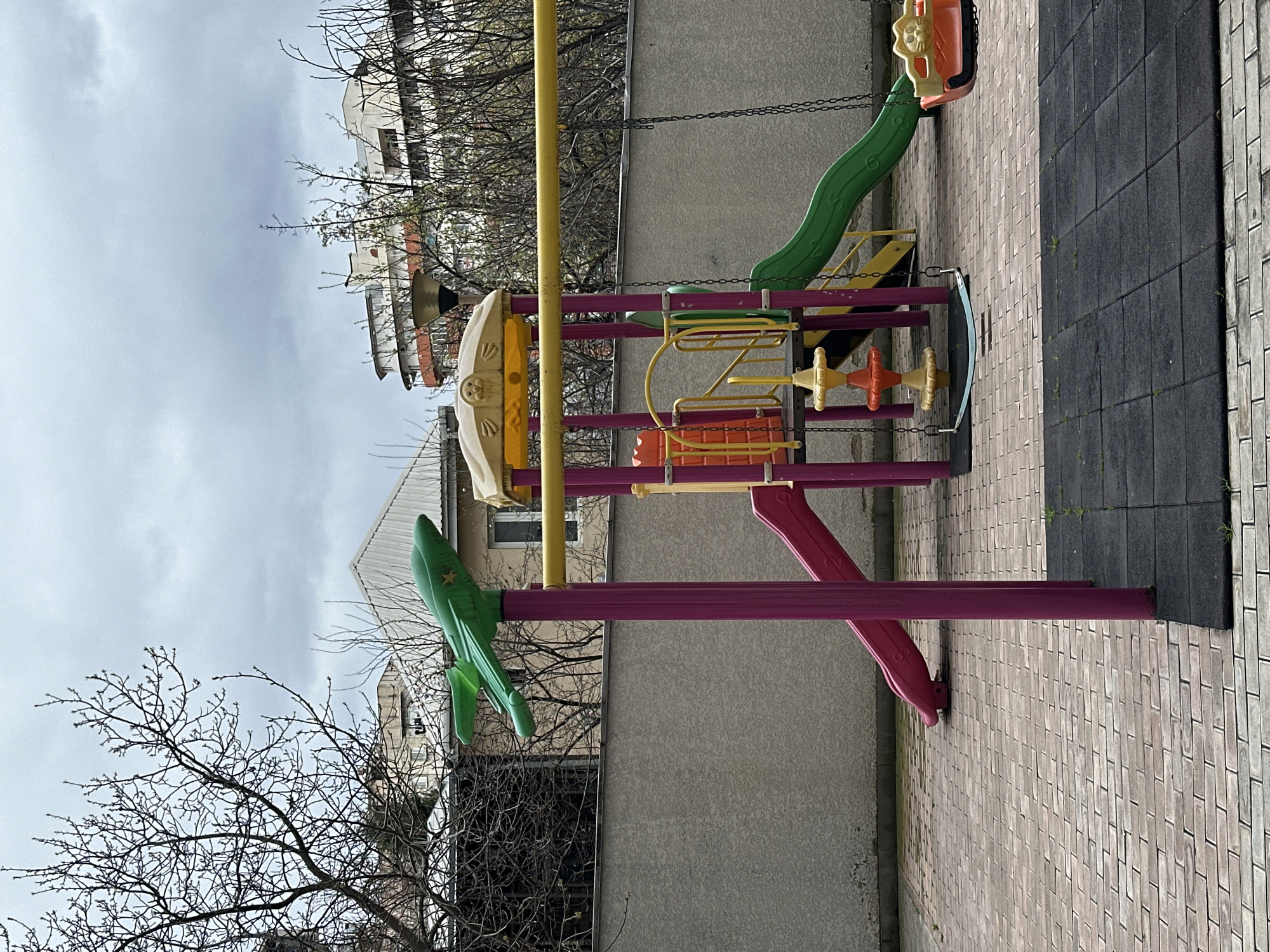 The image depicts an empty outdoor children's playground located in a residential area of Tbilisi, Georgia, under an overcast sky. The foreground features light-colored rectangular pavers that transition to dark gray rubber safety mats beneath the play equipment. Small sprouts of green vegetation are visible through the paving and mats.
The primary subject is a multi-component playground structure constructed from brightly colored plastics and metals. It consists of magenta support poles, yellow horizontal bars, a pink slide, and a green slide. Two swings are present: one with a blue flat seat and another with an orange toddler seat shaped like a cat's head. A green airplane-shaped ornament with a yellow star is mounted atop a magenta pole on the left side of the structure. A yellow canopy, featuring a sculpted face, crowns a section of the structure, alongside a multi-tiered orange and yellow play element.
Behind the playground, a textured light gray concrete wall acts as a boundary. Beyond the wall and numerous bare deciduous trees, various residential buildings are visible. These include a structure with a white sloped roof and a larger, multi-story building characterized by a rounded facade and terracotta-colored elements. A dark-colored street lamp is positioned above the playground. No individuals or active events are present within the scene.