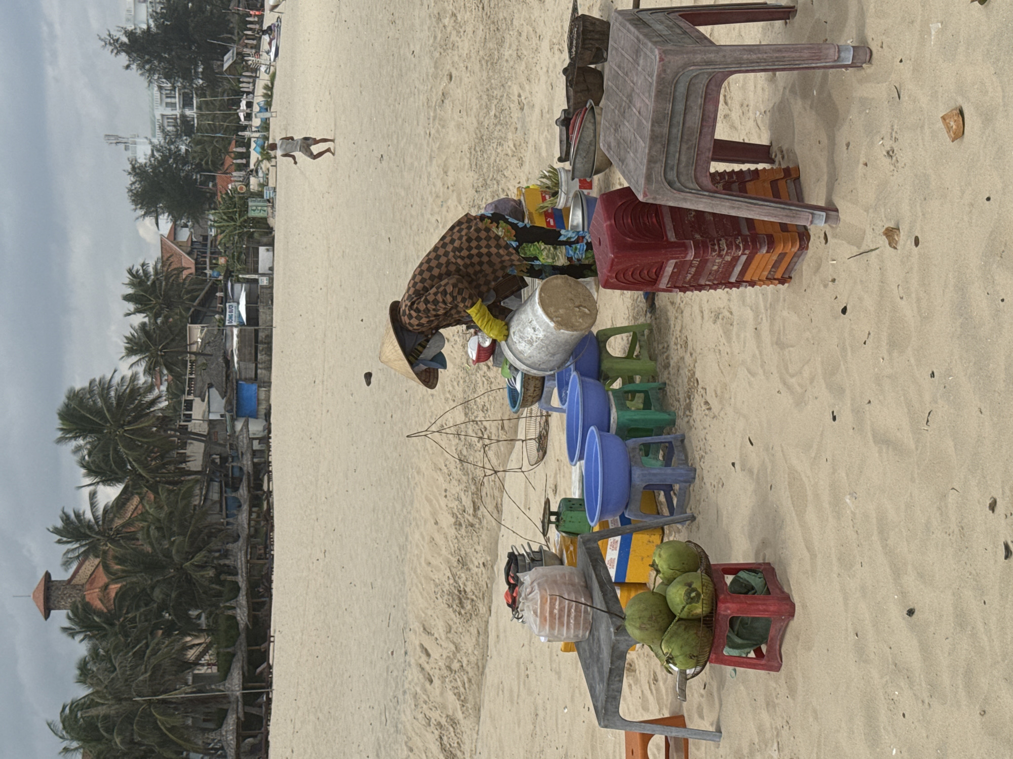 A person wearing a conical hat, yellow gloves, and a checkered shirt is standing on a sandy beach in Mui Ne, Vietnam, next to a table laden with coconuts, bowls, and containers. The person is holding a large metal bucket and appears to be in the process of pouring its contents. Behind the table are stacks of colorful plastic stools and various fishing or food preparation equipment. In the background, a resort-like building with a distinctive orange roof is visible amidst palm trees, and other beachgoers can be seen in the distance. The overall scene suggests a bustling marketplace or a vendor preparing for business on a warm, overcast day.