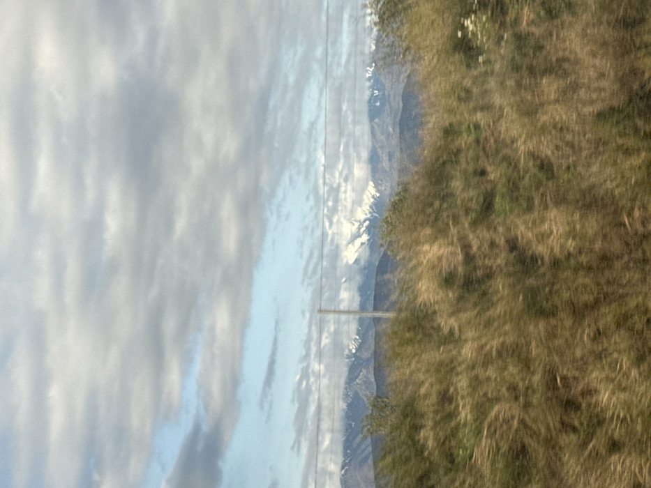 This image depicts a natural landscape scene, likely captured from a moving perspective in the vicinity of Canohès, France. The foreground is dominated by a blurred field of dry, tall brownish-green grasses and some greener vegetation, with a few small white floral clusters visible on the right. A wire fence with a single prominent vertical metal post spans horizontally across the midground. Beyond the fence, a distant mountain range is visible, characterized by snow-capped peaks and partially obscured by atmospheric haze or clouds. The sky above is largely overcast with grey and white clouds, interspersed with clear patches of blue, particularly towards the upper left. No individuals or specific actions are discernible within the frame. The overall environment suggests a rural or semi-rural setting under natural daylight conditions.