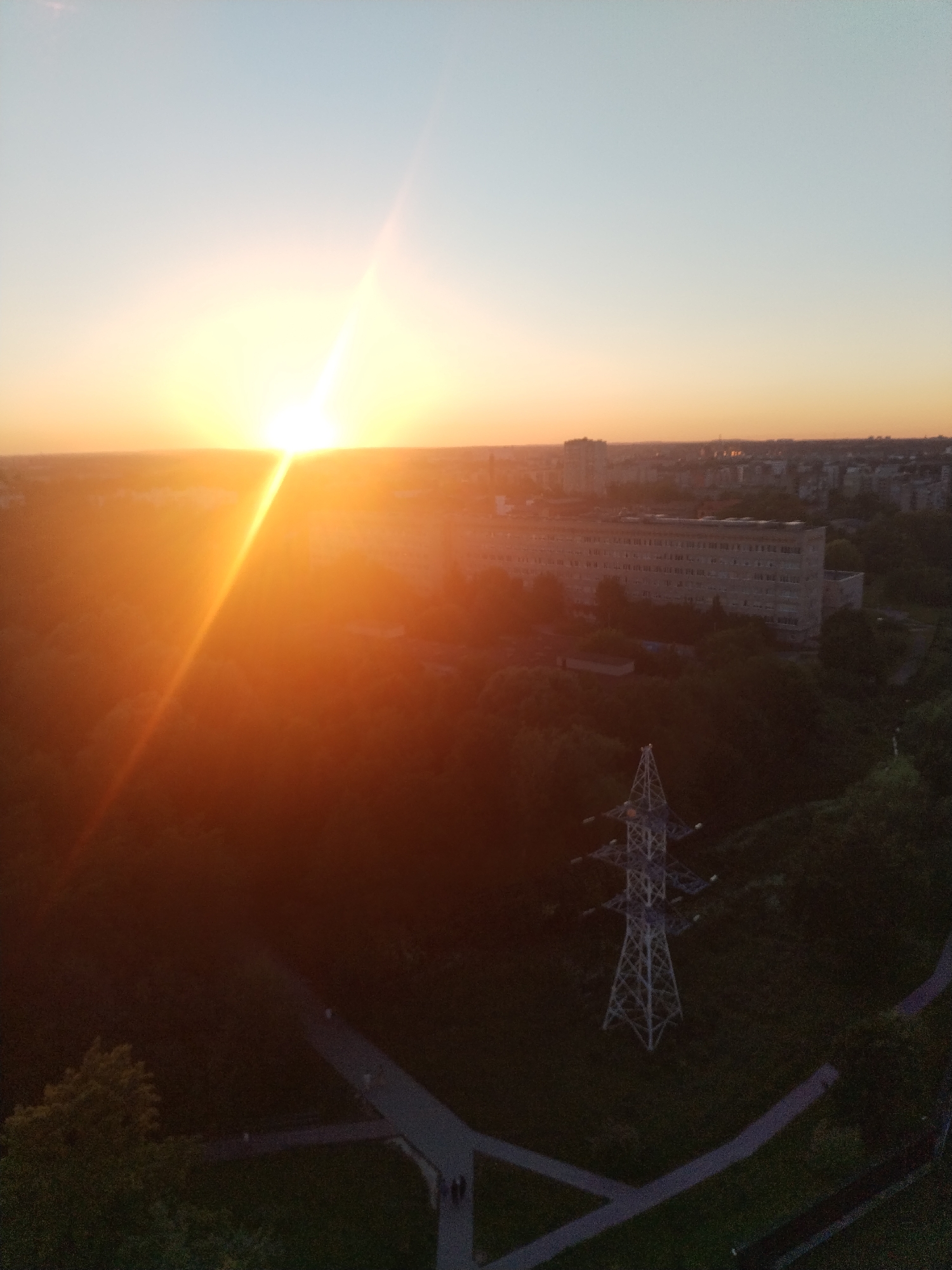 This aerial photograph depicts a sunset over a city, likely Kaliningrad, Russia. The sun, a brilliant white orb with orange rays, is positioned low on the horizon, casting a warm glow across the sky. Below the sun, a cityscape unfolds, characterized by a cluster of buildings with a large, rectangular structure dominating the mid-ground. The urban landscape is interspersed with lush green trees, suggesting a park or wooded area within the city. A prominent latticed power pylon stands in the foreground, its geometric structure contrasting with the organic shapes of the foliage. Winding paths, possibly pedestrian walkways, are visible, with a few individuals appearing as small figures, their actions indiscernible from this distance. The overall scene conveys a sense of urban tranquility as day transitions into evening.