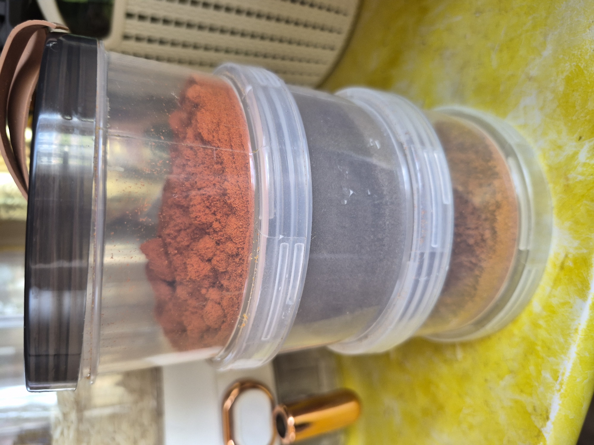 A close-up shot displays a stack of three transparent plastic cylindrical containers lying horizontally on a vibrant yellow surface. The topmost container, which also has a dark gray lid and a brown strap or handle attached, holds a substantial amount of reddish-orange powdered substance, possibly paprika or chili powder. The middle container is filled with a dark gray or black powder, which could be ground coffee or a dark spice blend. The bottom container contains a lighter orange or brownish powder, potentially turmeric or cumin.
The containers are designed to stack, evidenced by the ribbed edges where they connect. They rest on a brightly lit, irregularly patterned yellow surface, possibly a countertop or tablecloth with a marbled or textured finish. In the background, a light-colored, possibly slatted or woven, object is visible, suggesting an indoor setting during daylight hours. A portion of a golden or copper-colored object, perhaps a measuring spoon or part of a kitchen utensil, is also visible in the lower foreground. There are no people visible, no specific activity taking place, and no discernible text on any of the objects. The location context provided is Yerevan, Armenia, though no visual cues in the image explicitly confirm this.