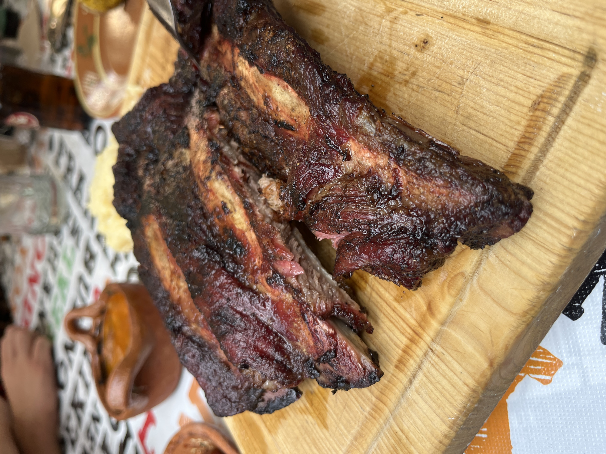 A close-up shot shows two pieces of heavily charred meat, likely ribs, resting on a light-colored wooden cutting board. The meat has a dark, almost black exterior with some visible red meat underneath. The cutting board is plain with visible wood grain. The setting appears to be a dining area, with blurry objects suggesting dishes and cutlery visible in the background. A tablecloth with a pattern of black and white geometric shapes is also present in the lower right corner.