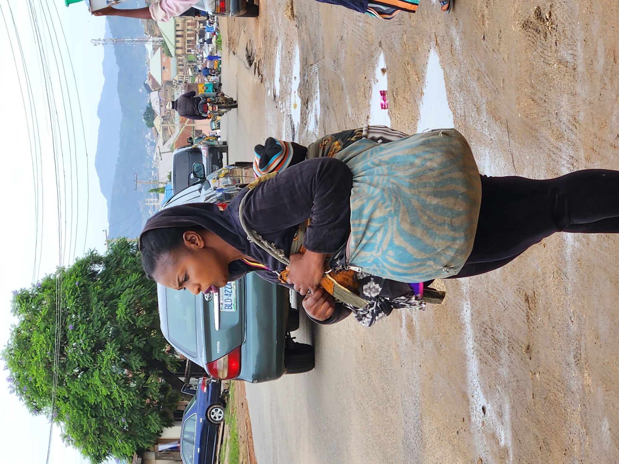 A woman with dark skin and dark hair, wearing a dark hooded jacket over a patterned cloth and black leggings, stands on a dirt road in what appears to be a city. She is carrying a baby wrapped in a patterned cloth on her back, and a large, light blue and tan patterned bag over her left shoulder. Her head is turned to her right, and her mouth is open as if speaking. The road is partially covered in puddles of water, indicating recent rain. In the background, a green Toyota sedan with a license plate reading "BLD 422A" is parked on the left side of the road. Further in the background, there are other vehicles, buildings, a tall tree with green foliage and purple flowers, and a hazy mountain range. The sky is overcast.