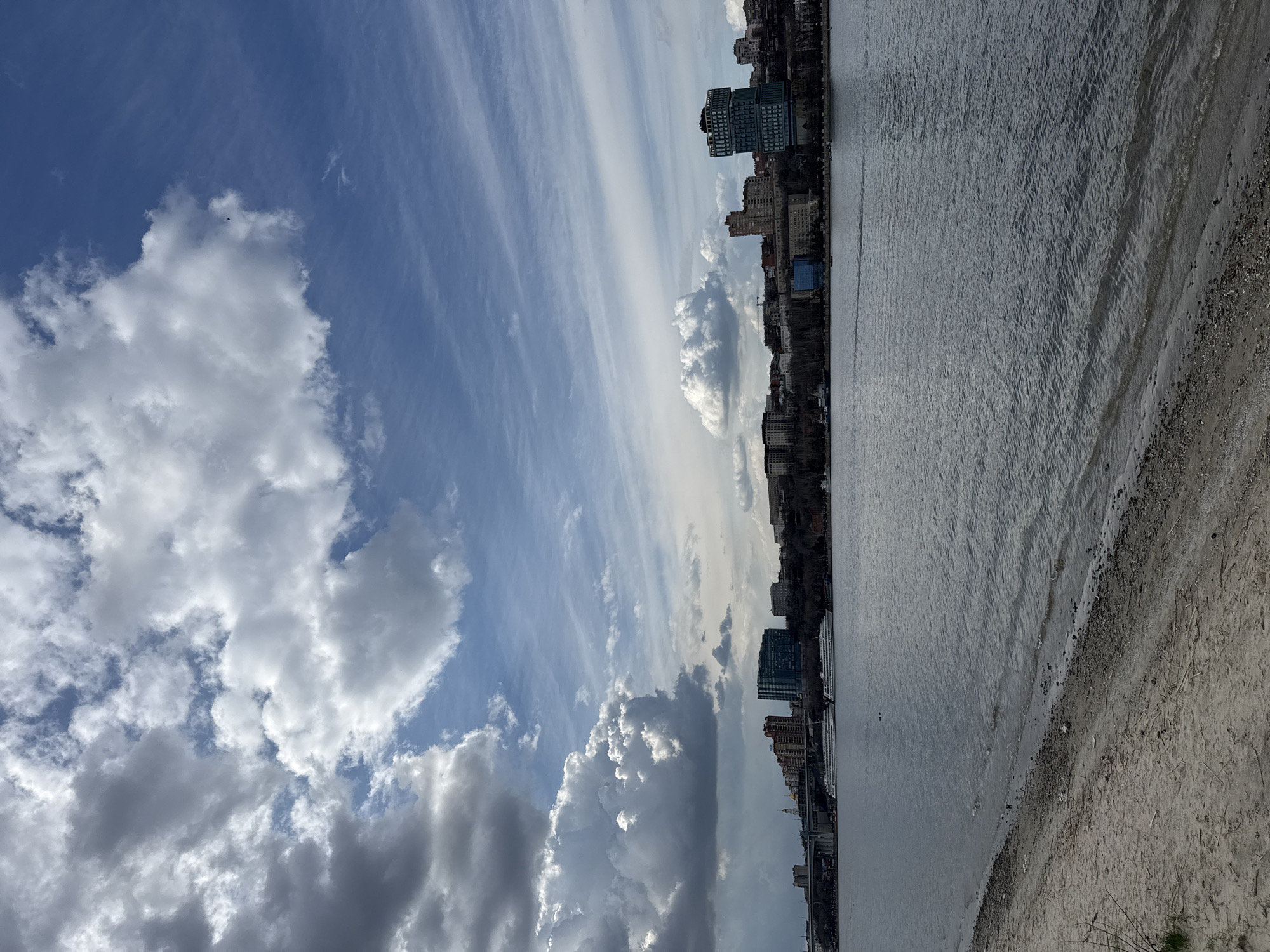 The image shows the Don River in Rostov-on-Don, Russia, with a view of the city skyline along the bank. The sky is mostly blue, interspersed with both scattered puffy cumulus clouds and long bands of cirrus clouds. The river's surface is slightly rippled, suggesting a gentle breeze. A narrow strip of beach runs along the river's edge in the foreground, composed of sand and small pebbles. Along the riverbank, there are several mid-rise and high-rise buildings visible, defining the cityscape. The buildings vary in architectural style and appear to be a mix of residential and commercial properties.