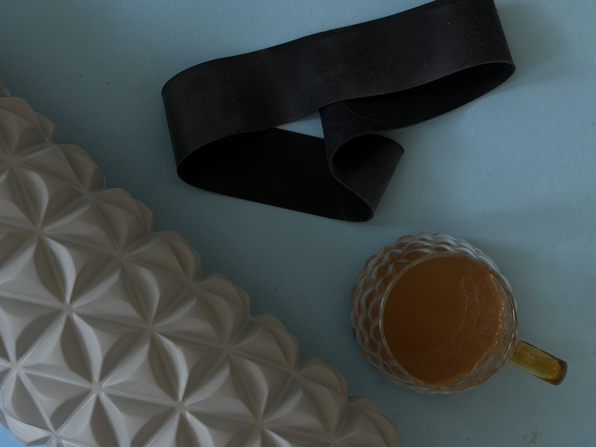 The image shows a blue surface with a foam roller, a black resistance band, and a glass mug with a brown liquid. The foam roller is white and has a textured surface with geometric patterns. The black resistance band is partially coiled, and the mug has a handle. The scene likely takes place indoors. The location in Izhevsk, Russia is not discernible from the visual information.
