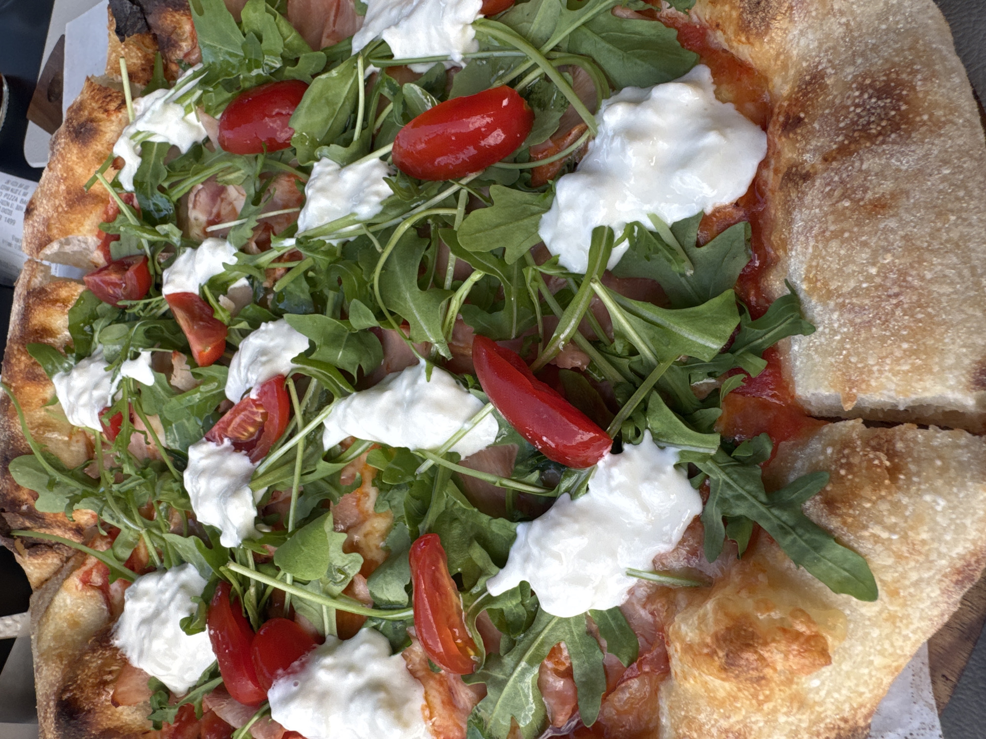 The image depicts a close-up of a pizza with a thick, airy crust. The crust is golden brown with some charred spots. Toppings include arugula, halved cherry tomatoes, and dollops of soft white cheese, likely burrata or a similar creamy cheese. There are also slices of prosciutto visible under the arugula. Based on the presence of arugula, prosciutto and burrata, the pizza is likely served at an upscale establishment in the city of Budva, Montenegro. No people or interactions are visible in the scene.
