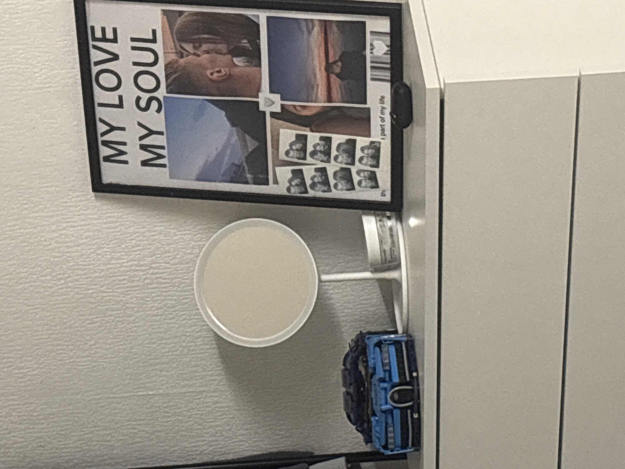 The image captures an indoor scene, likely a personal room, showcasing a framed picture and several objects arranged on a surface next to a cabinet. The primary subject is a large, black-framed picture or collage hanging on a textured, light-colored wall.
The framed picture is a collection of photos and text. On the left side, prominent black text reads "MY LOVE MY SOUL" stacked vertically. The right side features several images: a close-up photo of a couple kissing, a landscape shot with a person standing, and multiple small, photobooth-style strips showing a couple in various poses. Near the bottom right of the collage, small text reads "I part of my life," and there's a barcode-like pattern in the top right.
Below the framed picture, on a shelf or surface, sits a small, circular mirror with a white frame and stand. Next to the mirror, there is a small blue and black toy vehicle, possibly a Lego car or similar model. To the right of these objects is a light-colored, possibly white or off-white, two-door cabinet or wardrobe. A small black object, possibly a hook or sensor, is attached to the side of the cabinet, aligning with the framed picture. The wall has a distinct, subtle textured pattern. The overall impression is that of a cozy and personalized space.
Given the context, the location is Saint Petersburg, Russia.