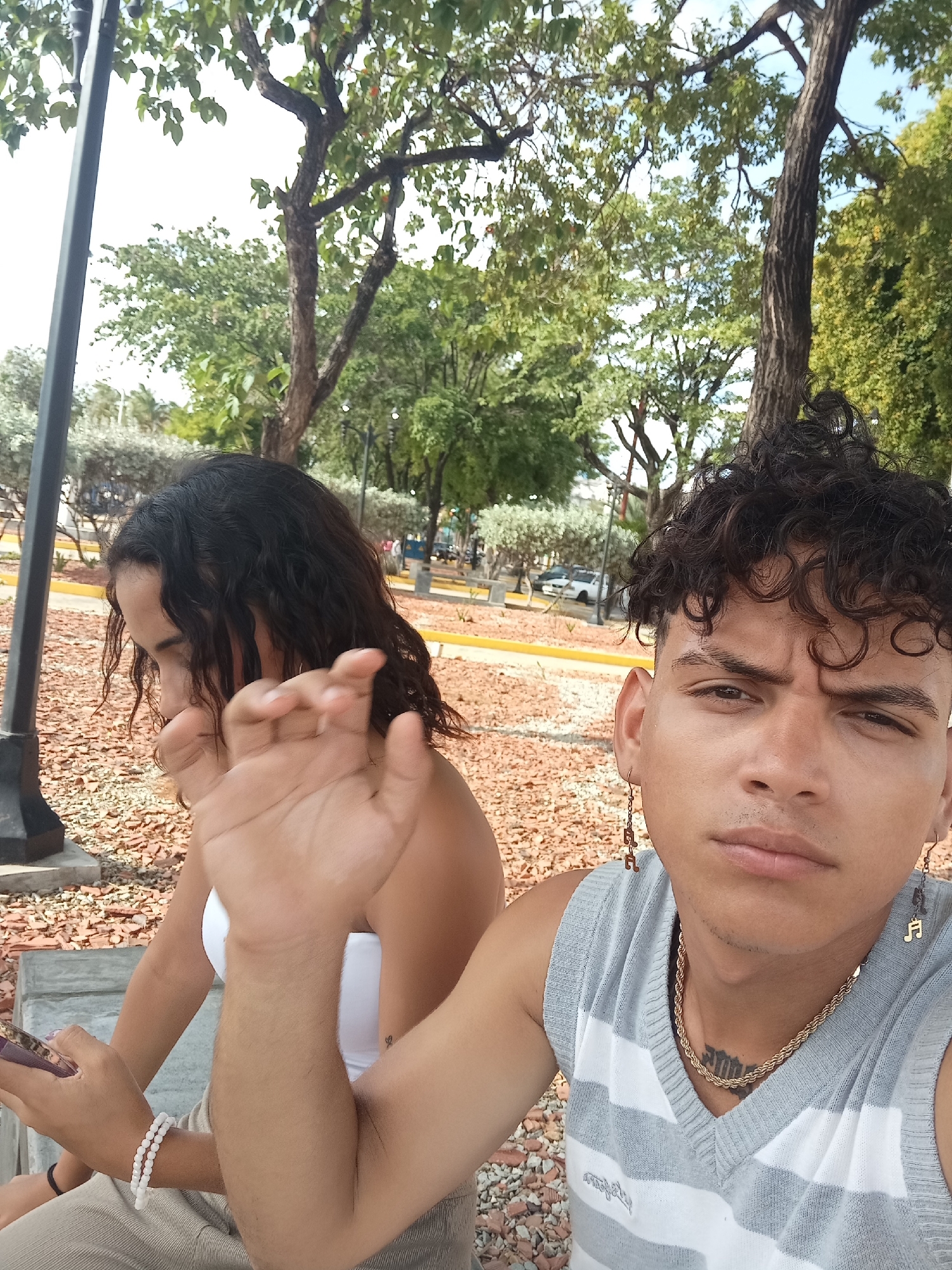 Two young adults are seated on a concrete bench outdoors. The person on the right, a male with curly dark hair, is in the foreground, facing the camera. He is wearing a grey and white striped v-neck sweater vest, a gold chain necklace, and earrings shaped like musical notes. He has a tattoo on his chest. The person on the left, a female with long, wavy dark hair, is partially visible behind him, looking to her left. She is wearing a white strapless top and a bracelet made of white beads. She is holding a phone in her left hand. The background shows a park-like setting with trees, bushes, and a pathway. The ground is covered in reddish-brown gravel. In the distance, vehicles and buildings can be seen. The overall lighting suggests it is daytime.