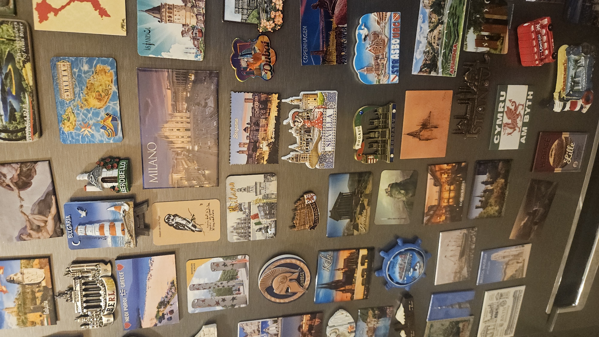 The image shows a collection of souvenir magnets attached to a metallic surface, likely a refrigerator door. The magnets depict various landmarks, cityscapes, and cultural symbols from different locations around the world. Some of the recognizable locations include Dubai, Copenhagen, Strasbourg, Yerevan (Armenia), Istanbul, London, Lille, Milan, Bergamo, Seville, and Budapest. Several magnets represent Bulgaria, including one depicting the meanders of the Arda River and a lighthouse with the inscription "Bulgaria." There's also a magnet specifically labeled "Plovdiv, Bulgaria." Other notable magnets include those representing Greece, Malta, and Wales. The arrangement appears somewhat random, covering a large portion of the metallic surface.