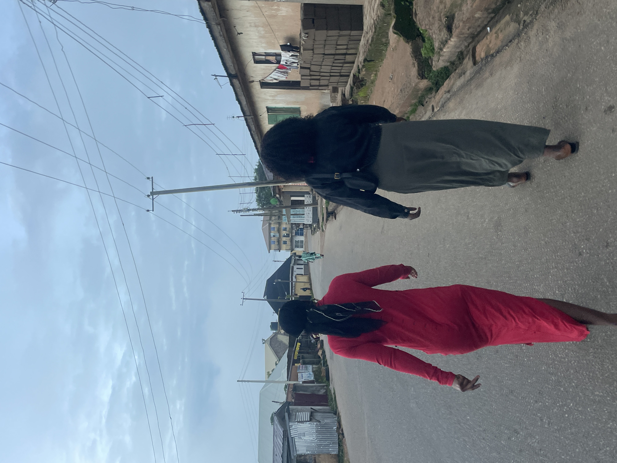 Two individuals are walking away from the camera down a street in what appears to be the city of Jos, Nigeria. The person in front is wearing a long, bright red dress and has their hair pulled back. The person behind them is wearing a dark jacket over a long, olive-green skirt and has voluminous dark hair. The street is paved with asphalt and lined with buildings on either side, some of which are constructed from corrugated metal and others from brick. There are power lines overhead, supported by wooden poles. Stacks of bricks are visible outside one of the buildings, and laundry is hanging to dry. The sky is overcast with a light blue hue. The scene suggests a residential or commercial area in an urban setting.