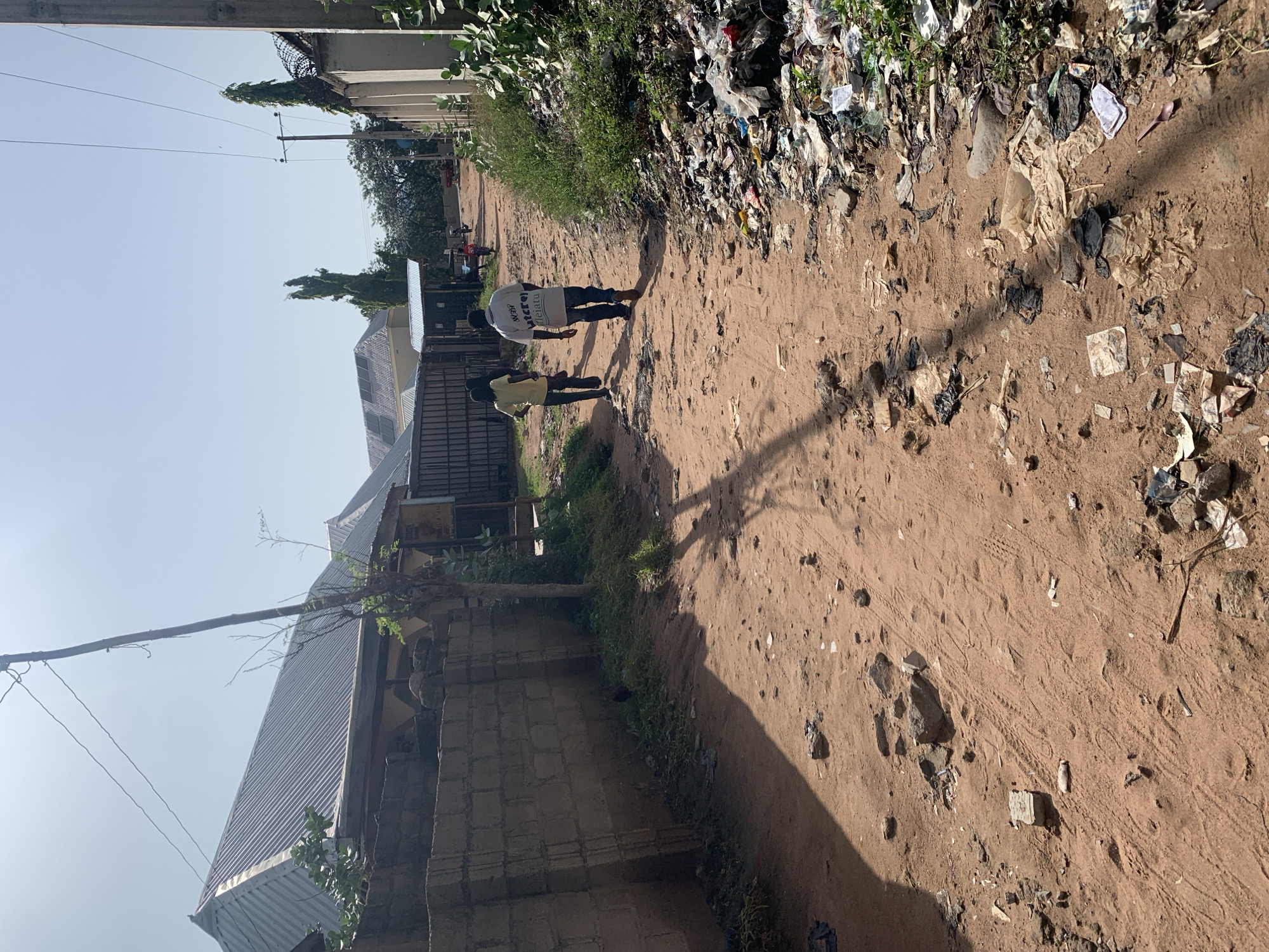The image shows an unpaved dirt road in Jalingo, Nigeria. The road is lined with buildings and fences on both sides. The ground is covered in red dirt, rocks, and scattered debris, including plastic bags, wrappers, and other refuse. On the right side of the road, a large pile of garbage is visible.
Two individuals are walking away from the camera on the road. The person in front is wearing a yellow t-shirt and dark pants. The person behind them is wearing a white t-shirt with text on the back and dark pants. In the background, there are more buildings, some with corrugated metal roofs, and several trees. Overhead, power lines stretch across the sky. The lighting suggests it is daytime, possibly late morning or early afternoon, with the sun casting shadows.