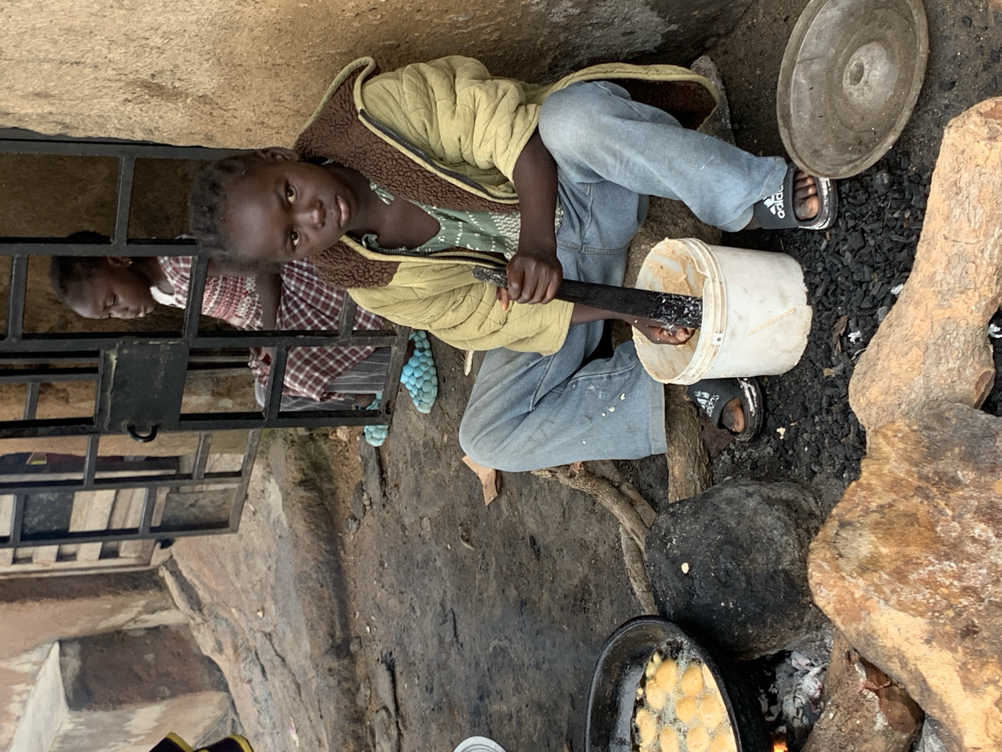 In Jos, Nigeria, a young girl sits on the ground, actively engaged in preparing food. She is wearing a light-colored quilted jacket over a patterned green top and blue jeans. In her hands, she holds a dark wooden pestle, which she is using to grind ingredients in a white bucket. Her feet are clad in black slide sandals with white accents.
Adjacent to her, a black frying pan is suspended over a fire fueled by charcoal. The pan contains several golden-brown, fried food items immersed in hot oil. To the right of the bucket, a circular, textured, grey plate rests on the ground. The environment appears to be an outdoor setting, with a rough, dirt-covered ground and a weathered, beige wall in the background. Behind the girl, through a metal grate, another young girl can be seen in the background, wearing a red and white checkered dress. The overall scene depicts a moment of domestic activity and food preparation.