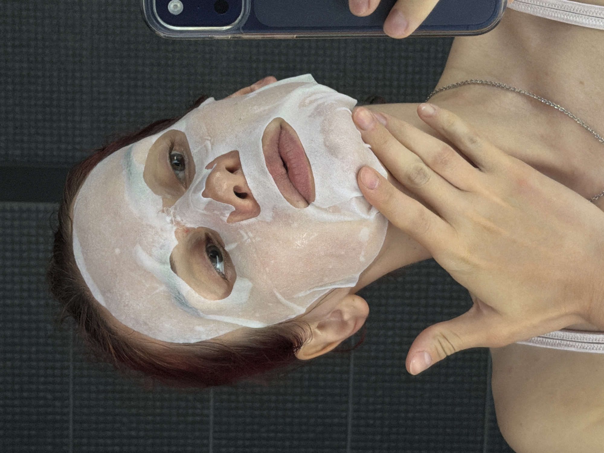 A woman lies with her head angled towards the upper-left corner of the frame, wearing a white sheet mask. Cutouts in the mask expose her eyes, nose, and lips. Her right hand rests on the mask, near her cheek. She holds a dark-colored smartphone with two rear-facing cameras in the upper-center. A silver necklace is visible on her neck, as is the strap of what appears to be a white bra. The background consists of gray tiles. No information suggests this image was taken in Chaikovsky, Russia.