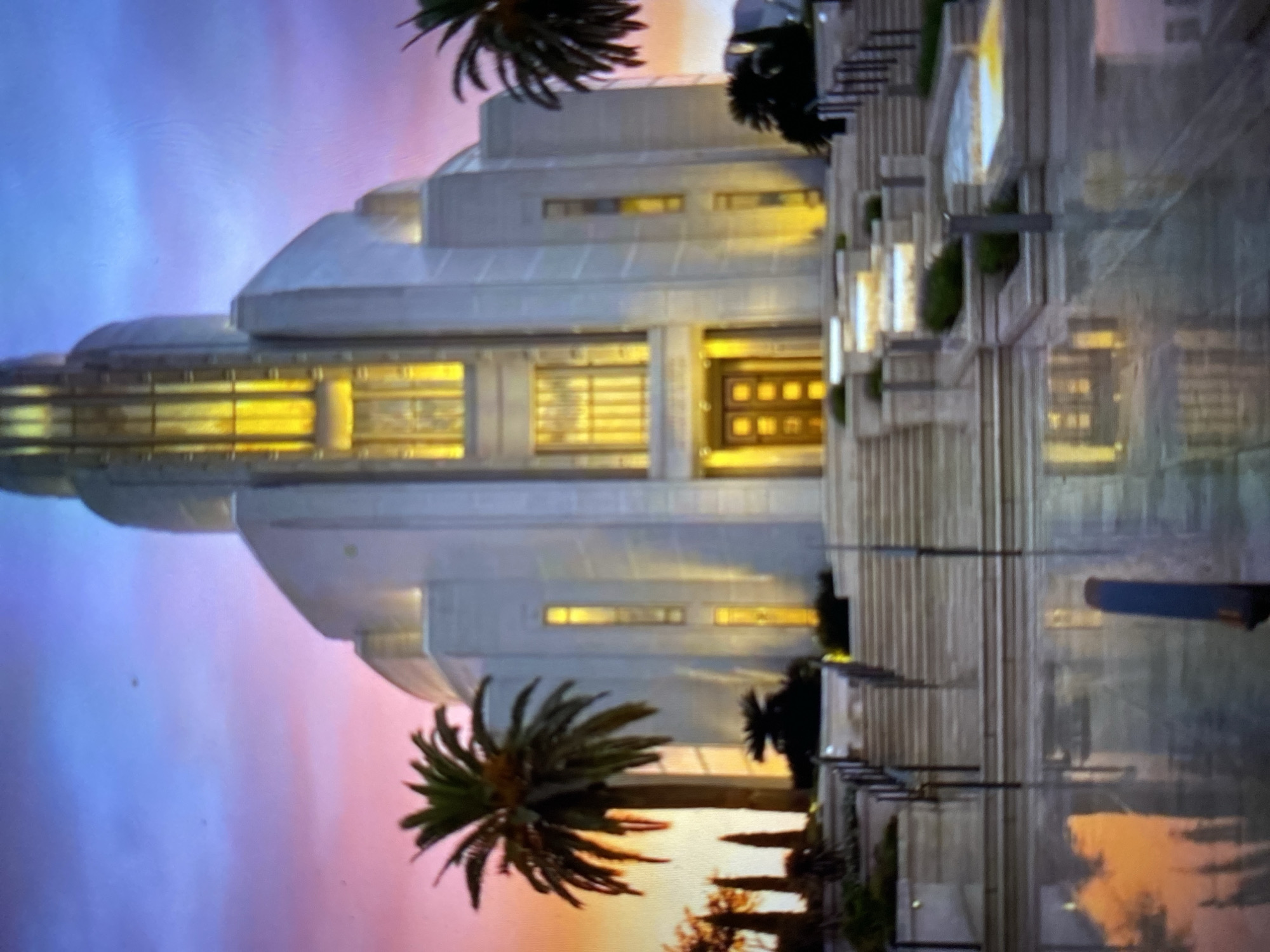 The image depicts a tall, white, multi-tiered building, likely a religious temple, illuminated from within and featuring a central door flanked by windows. Two palm trees frame the building, set against a colorful sunset sky that transitions from blue to pink. The temple’s front steps and surrounding walkways are visible, with reflective surfaces indicating moisture, possibly from recent rain. The image is presented sideways and appears to be a photograph of a scene in Tinaquillo, Venezuela.