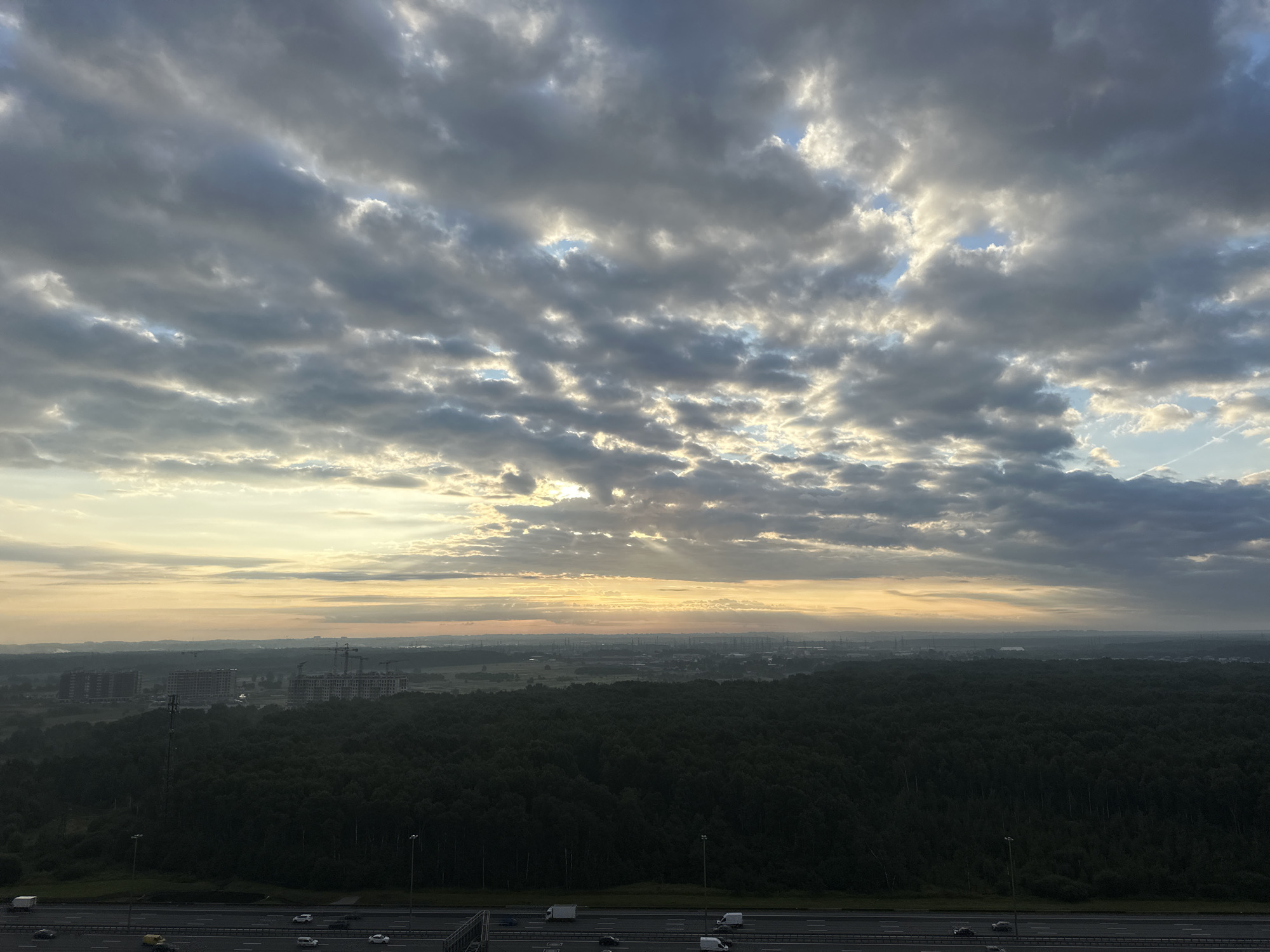 The image shows a wide-angle view of a cityscape and its surrounding environment, possibly at sunrise or sunset, given the warm light and dramatic cloud formations. In the foreground, a multi-lane highway is visible with several cars and trucks driving on it. Beyond the highway, a dense, dark forest stretches across the landscape. In the mid-ground, several large buildings under construction are apparent, with cranes visible amongst them. The background shows a hazy expanse with more distant structures and possibly industrial elements. The sky is filled with a mixture of dark, heavy clouds and patches of lighter, illuminated sky, suggesting changing weather conditions or the specific time of day. The overall impression is one of urban development encroaching on a natural landscape under a dynamic sky. The location is specified as Lednevo, Russia.