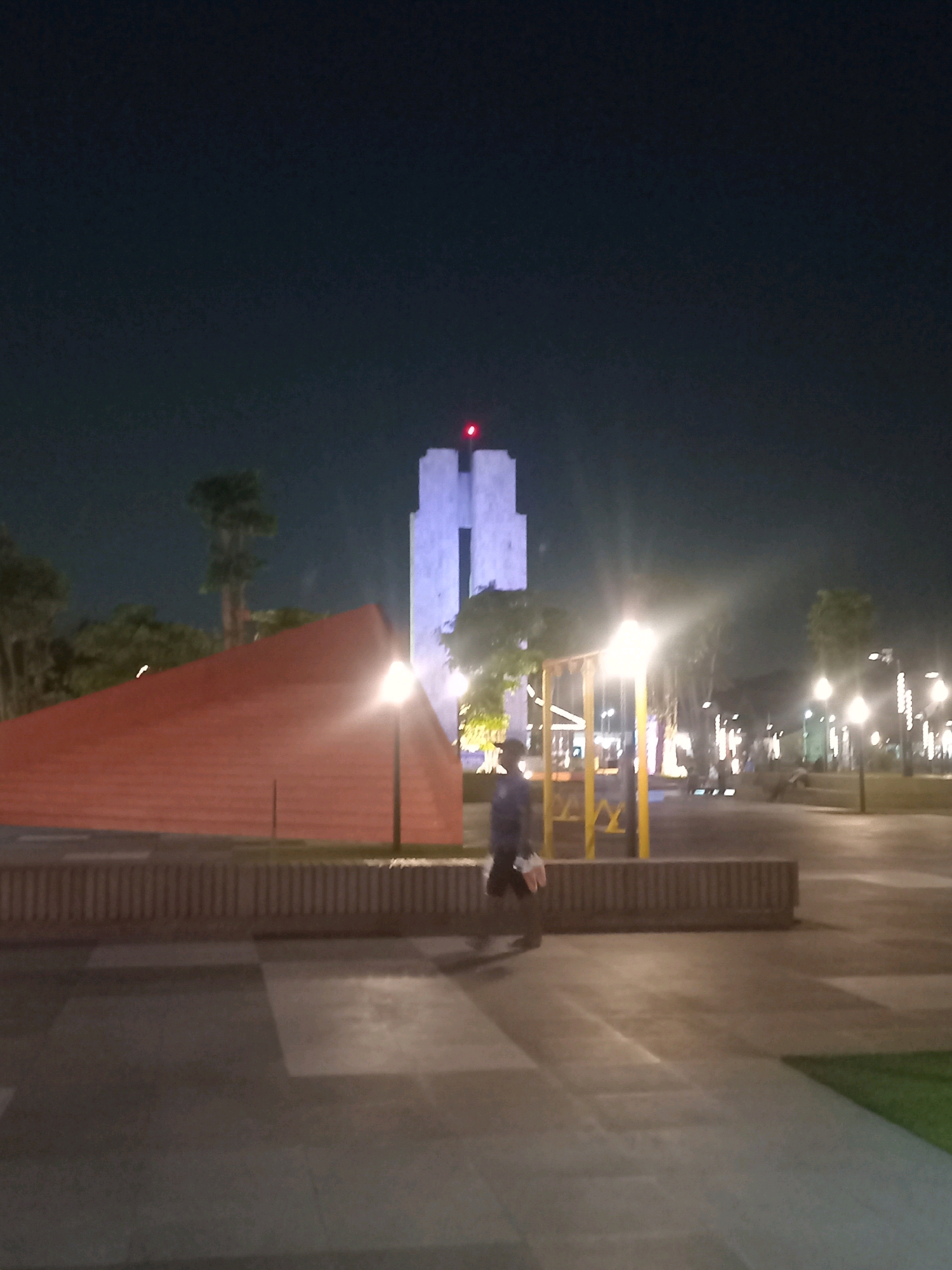 A nighttime, medium-shot, outdoor scene depicts a plaza in Sidoarjo, Indonesia. In the background, a tall, white, angular building with blue lighting and a red light on top stands against a dark sky. In the mid-ground, a person walks across a paved area. To the left of the person is a large, orange, triangular structure. To the right of the person is a yellow playground structure with lights. Scattered streetlights illuminate the paved plaza.