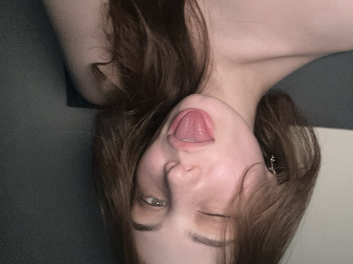 The scene depicts an indoor setting, contextually located in the city of Kommunarka, Russia. A single individual with fair skin and long, reddish-brown hair is centered in a close-up, chest-up view. The individual's left eye is closed in a wink, while the right eye is open, revealing a green iris. The tongue is fully extended from the mouth. A small, metallic, dangle earring is visible on the left ear. The shoulders and upper chest are bare. The background consists of a dark, undifferentiated surface on the right and a lighter, off-white surface on the upper left. Illumination appears dim and artificial.