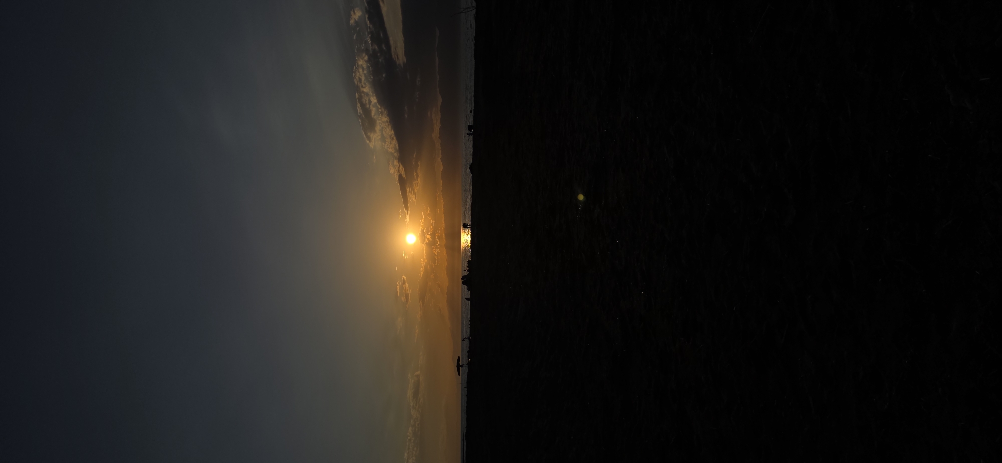 This is a vertical, full-frame photograph of a beach scene at sunset. The sun is low on the horizon, casting a golden glow and reflecting on the water. The sky transitions from a dark, deep blue at the top to a warm orange and yellow near the horizon. Clouds are scattered across the sky, some catching the sunlight.
In the middle ground, a silhouetted shoreline is visible, with faint mountain ranges in the distance. Along the beach, several small, dark figures are scattered, some appearing to be individuals or small groups of people. One figure is partially obscured by what could be a beach umbrella. The water's surface reflects the bright sun.
The foreground is almost entirely black, showing the dark sand of the beach. A faint, yellowish-green lens flare is visible in the lower middle section of the foreground. The overall mood is serene and tranquil, capturing the end of a day at the beach. The scene is located in Boz Beshik, Kyrgyzstan.
