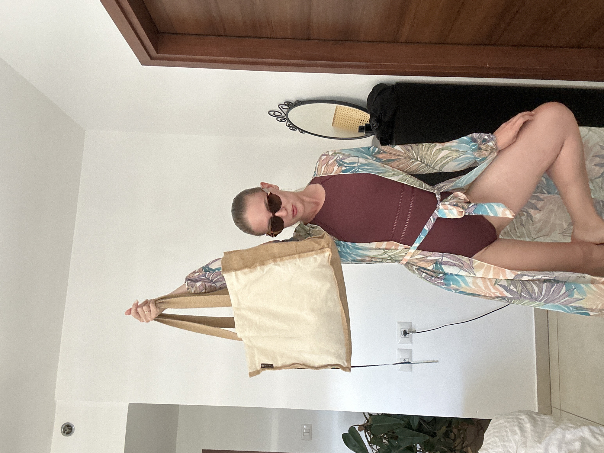 In an indoor setting, likely a room in Petah Tikva, Israel, a woman stands holding a large, light-colored tote bag with tan handles aloft. She is wearing a burgundy one-piece swimsuit and a sheer, long-sleeved robe with a tropical leaf print. The robe is cinched at the waist with a matching belt. The woman has her hair pulled back and is wearing sunglasses with brown frames. To her right, a dark cabinet stands against a white wall, topped with a small, oval mirror with an ornate black frame. Behind the cabinet, a closed wooden door is visible. In the background, a wall features electrical outlets and a dangling cable. To the left of the frame, a portion of a leafy green plant and a white, rumpled surface, possibly bedding or upholstery, are visible. The lighting appears to be natural, coming from an unseen source.