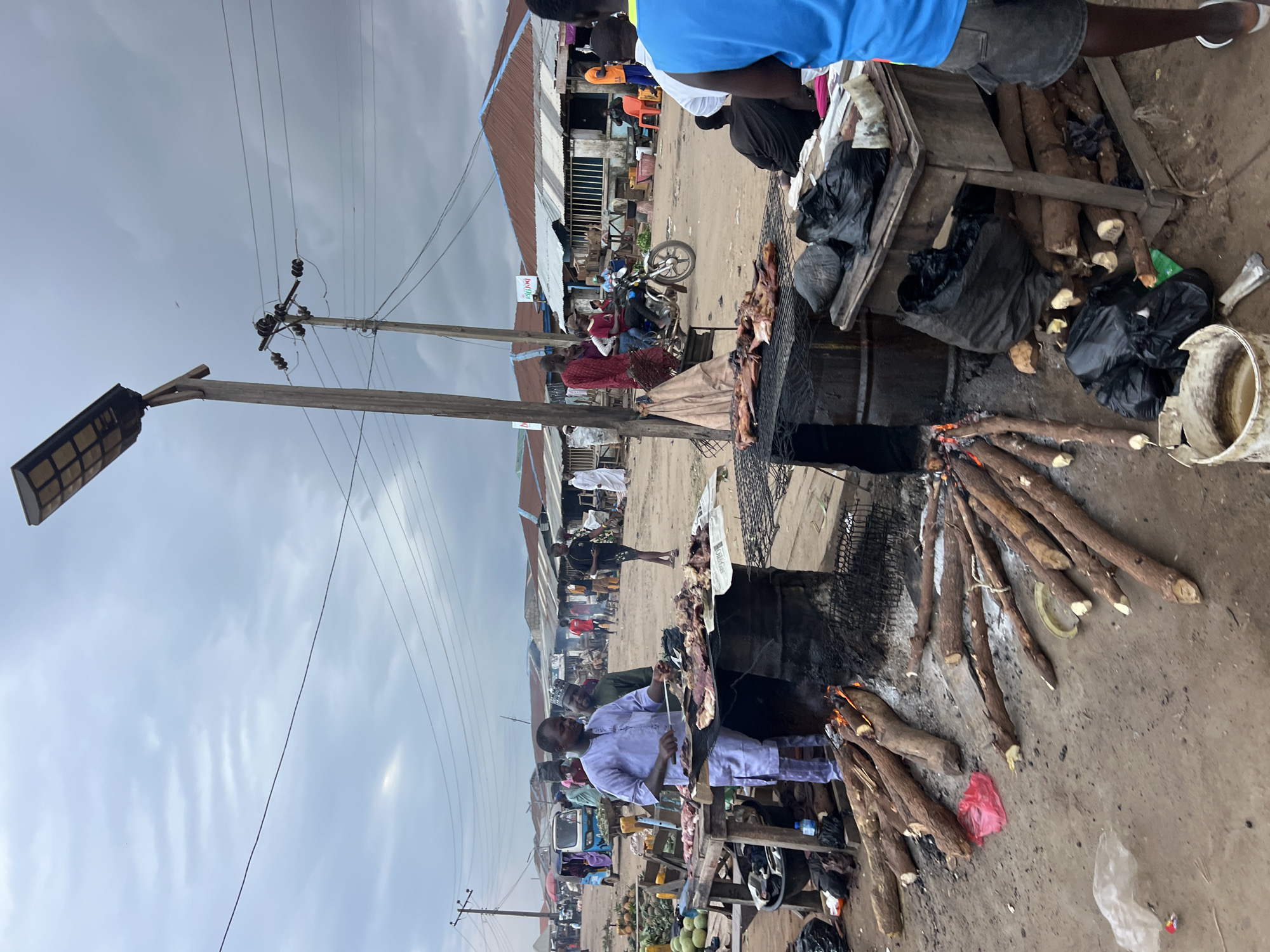 The image depicts an outdoor market scene in Oko Amakom, Nigeria. In the foreground, a man in a light purple traditional outfit is grilling meat on a makeshift barbecue, which consists of metal drums and a grate. Several pieces of raw meat are laid out on the grill. The fire beneath the grill is fueled by logs of wood. To the left of the grilling station, a pile of raw tubers, likely cassava, is visible. Various black plastic bags and some white plastic sheeting are scattered on the ground around the market stalls.
In the mid-ground, several other market stalls with corrugated metal roofs are visible, along with a few people engaged in market activities. A motorcycle with a rider and passenger is moving through the scene. In the background, more market stalls and structures extend into the distance. The sky is overcast with dramatic clouds. The overall impression is a bustling, everyday market scene in a Nigerian town.