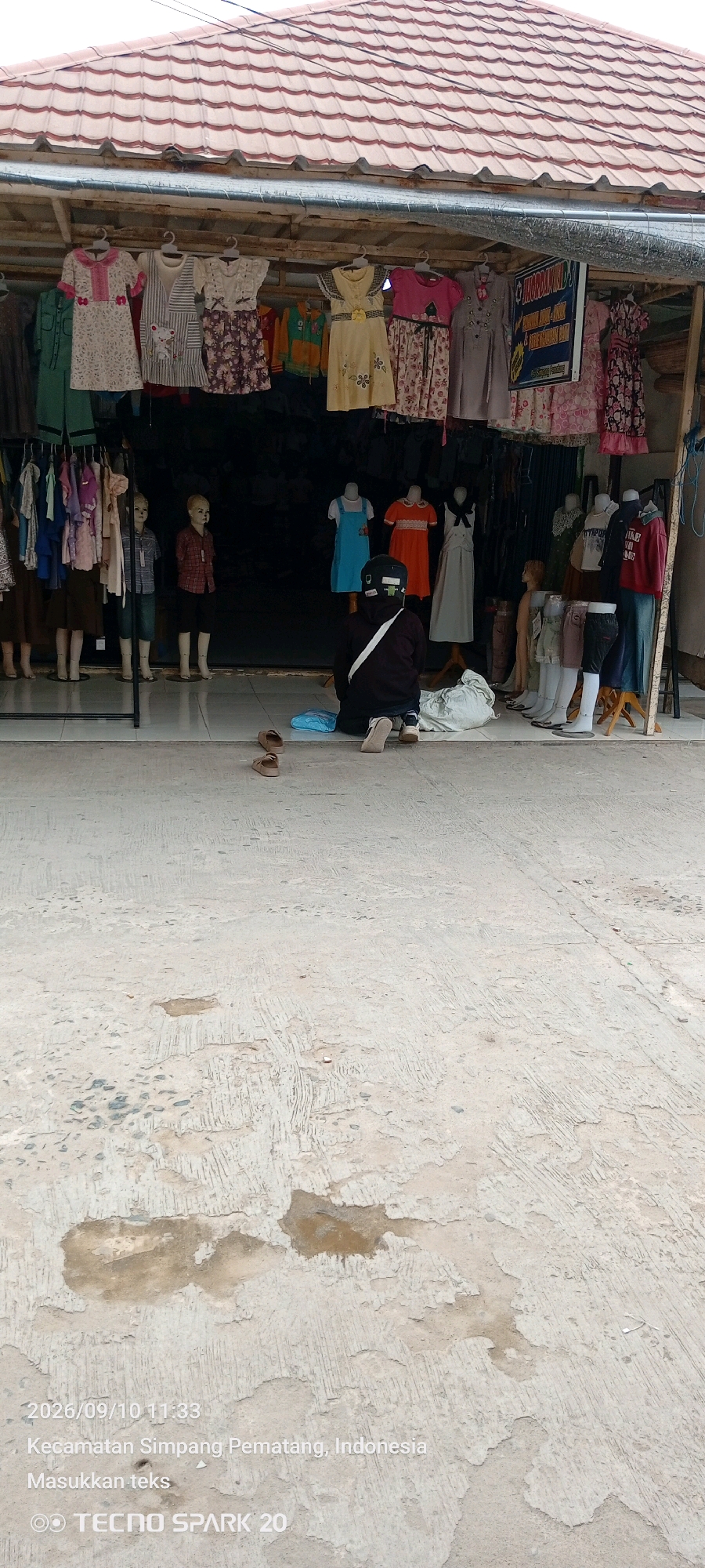 The image depicts a clothing stall located in the city of Simpang Pematang, Indonesia. The stall features a variety of clothing items, including dresses and outfits, displayed on hangers and mannequins. A person is kneeling in front of the stall, facing away from the camera, with a bag and a pair of sandals nearby. The ground in front of the stall is made of concrete with visible wear and patches of moisture. The overall scene suggests an outdoor market or a small shop with an open-front design. The timestamp and location information embedded in the image indicate the photo was taken on September 10, 2026, at 11:33 AM.