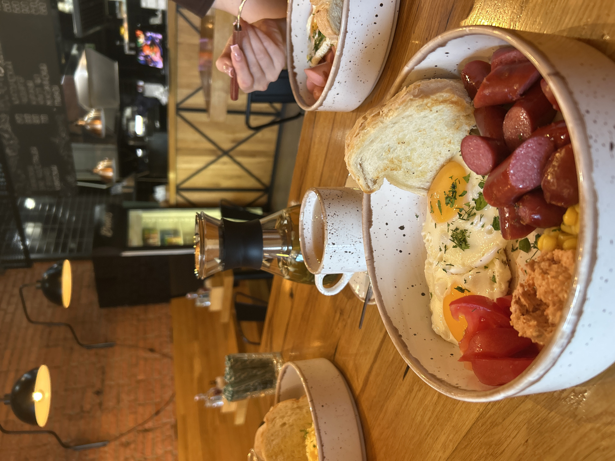 An overhead medium shot captures a wooden table in a dimly lit cafe, showcasing a hearty breakfast spread. In the foreground, a white ceramic bowl brims with a vibrant assortment of food: two fried eggs, sliced cherry tomatoes, a dollop of creamy orange spread, a scattering of corn kernels, and a pile of sliced, reddish-brown sausages. A piece of toasted bread rests on top. Adjacent to this bowl is a delicate white ceramic mug filled with amber-colored tea, resting on a saucer. Beside it, a glass carafe containing a light green liquid, possibly iced tea or a beverage, stands tall. In the background, another white ceramic bowl is partially visible, holding a similar breakfast dish. The cafe's rustic ambiance is evident in the exposed brick wall and wooden accents. The background is blurred, hinting at the cafe's interior with a counter and more patrons. A person's hands are visible on the right, holding utensils and interacting with their food. The scene evokes a sense of a relaxed and satisfying meal in a cozy cafe setting.